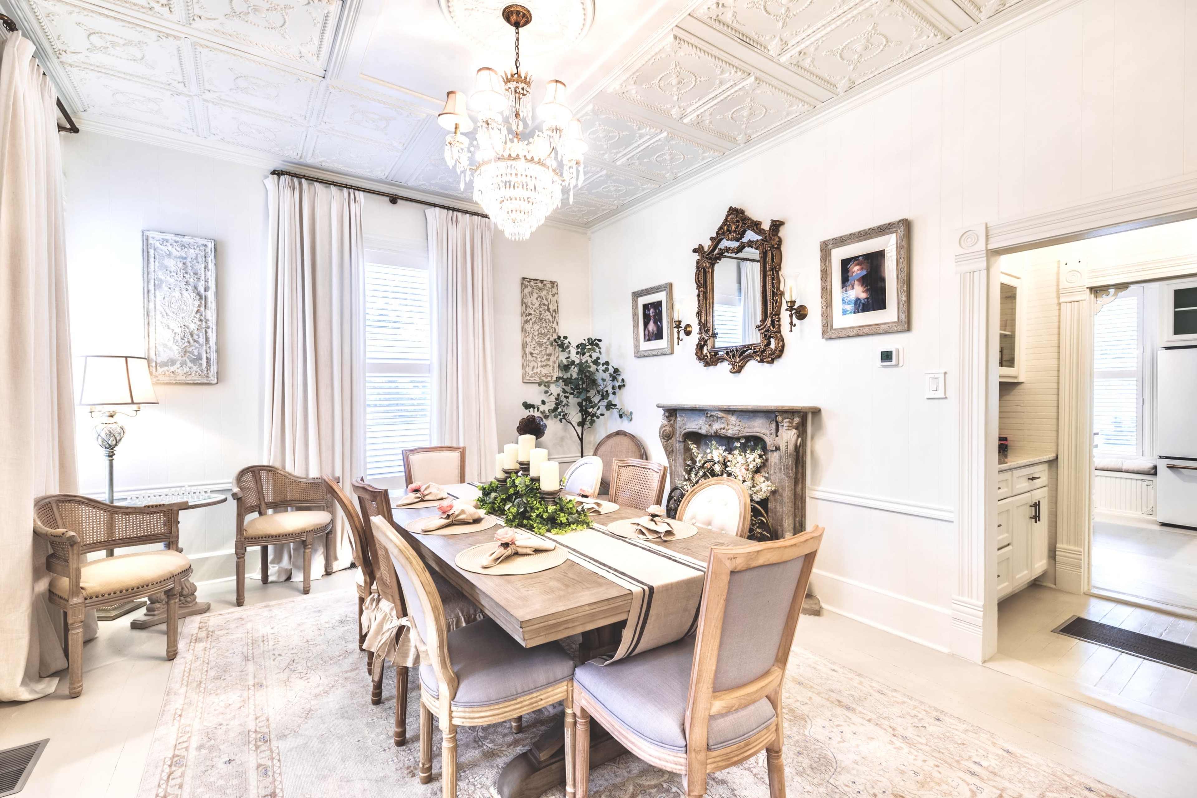 The image shows a bright dining room with a wooden table set for a meal, surrounded by upholstered chairs, a chandelier overhead, and decorative elements on the walls.
