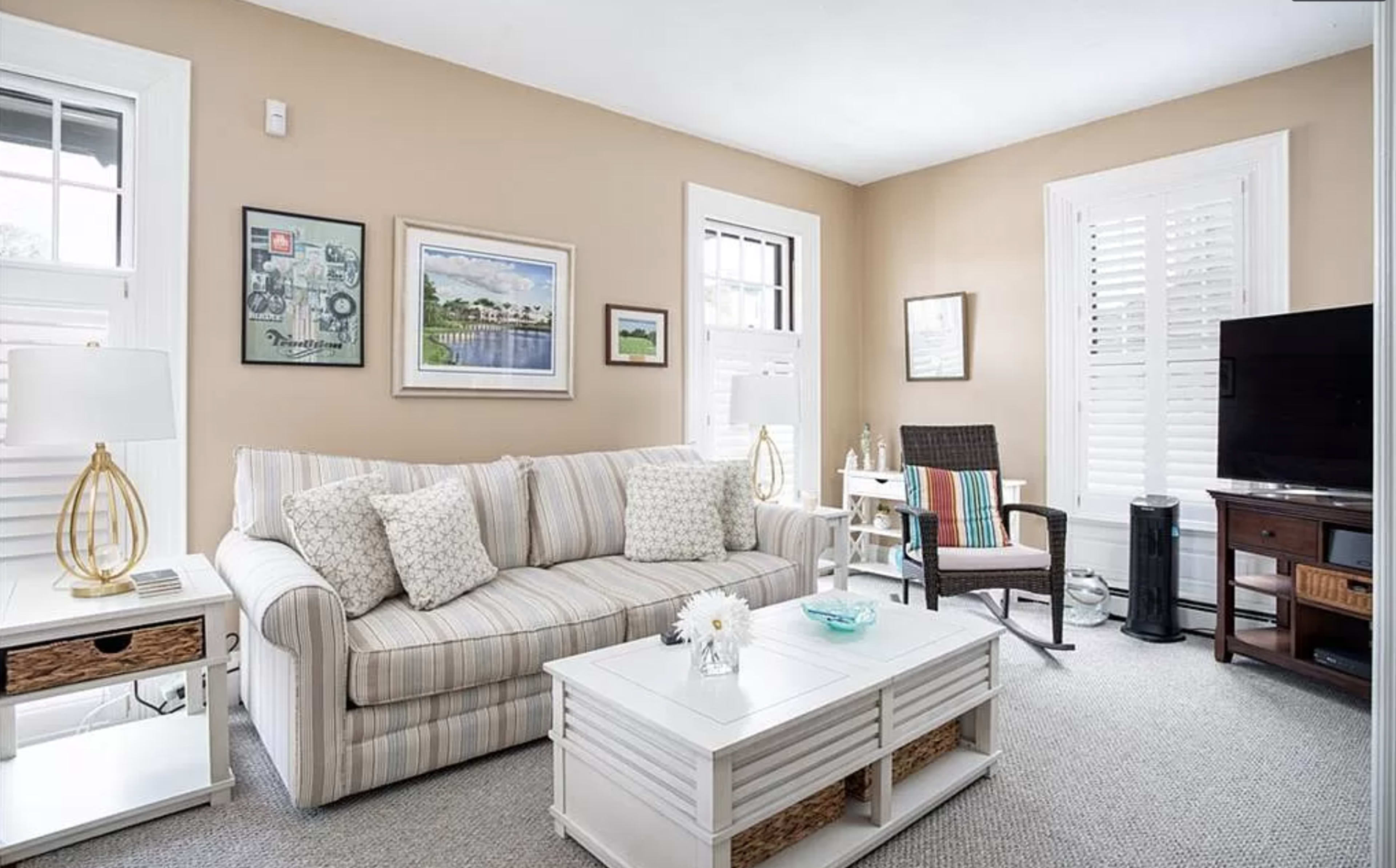 The room features a cozy living area with a striped sofa, a coffee table, and a television alongside a window with white shutters.