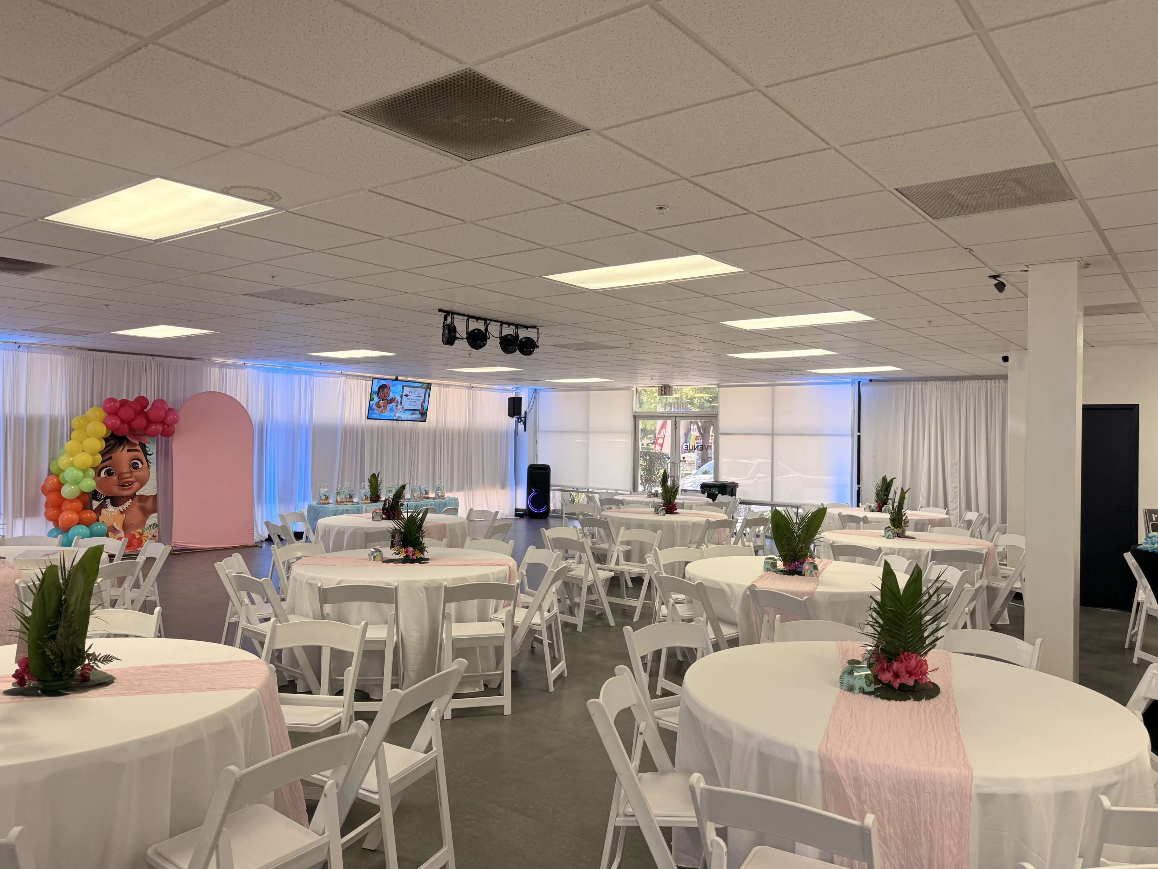 The image shows a spacious event room set up with round tables covered in white tablecloths, decorated with floral centerpieces and surrounded by white chairs.