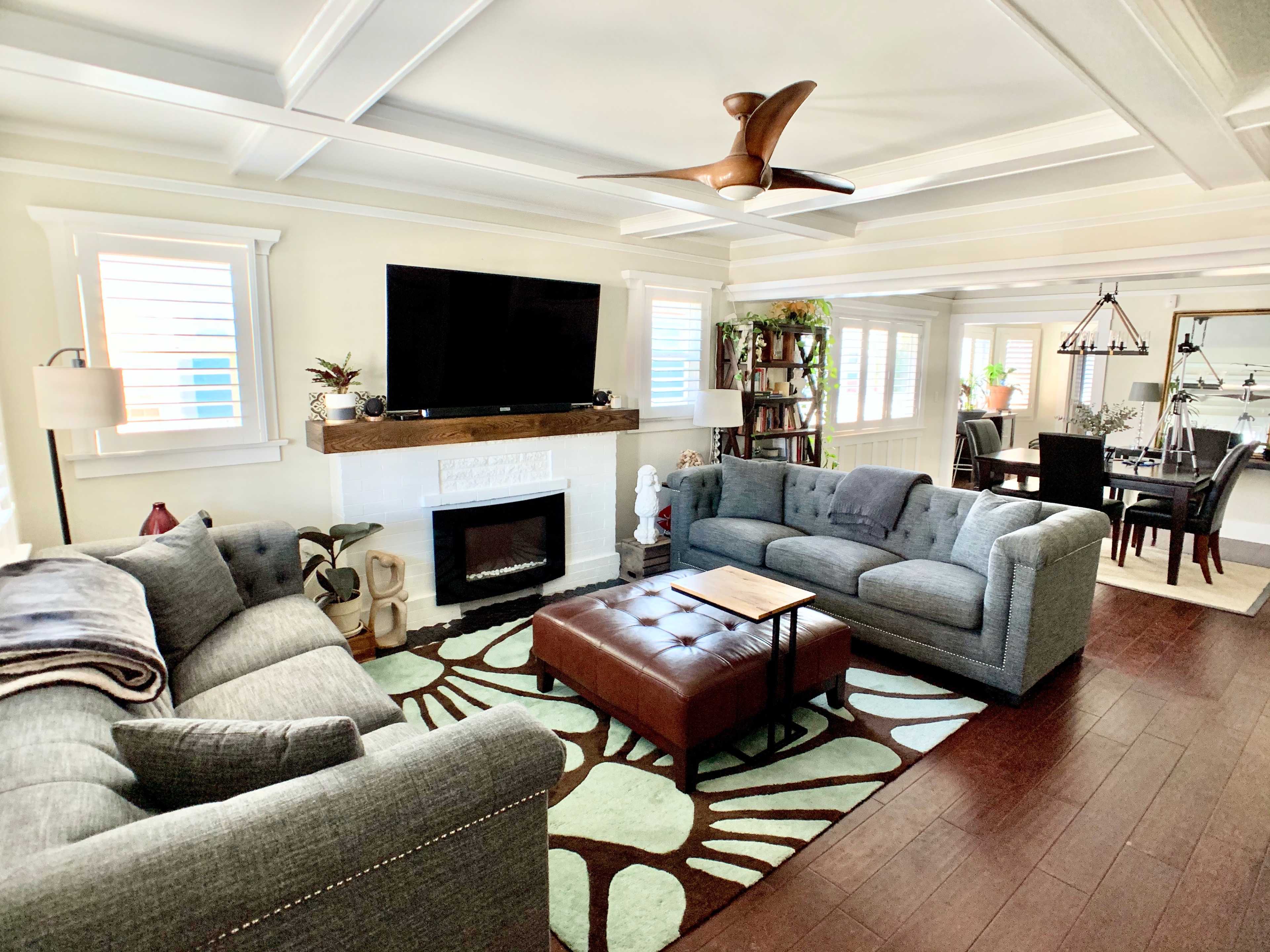 The living room features two gray sofas facing a coffee table, with a flat-screen TV mounted above a white fireplace, and a dining area visible in the background.