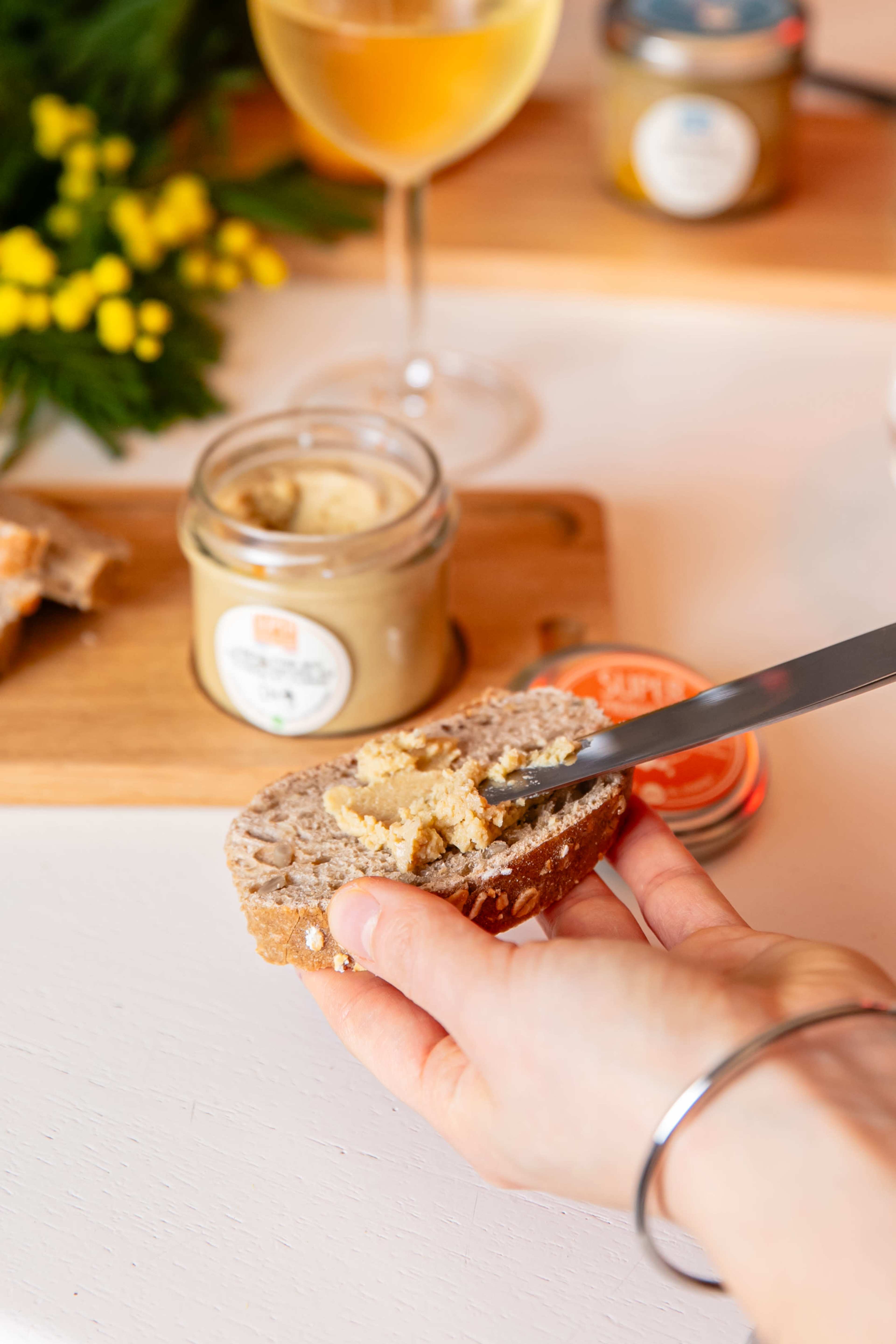 A hand spreads a creamy substance onto a slice of dark bread, with a jar of the substance and a glass of wine in the background.