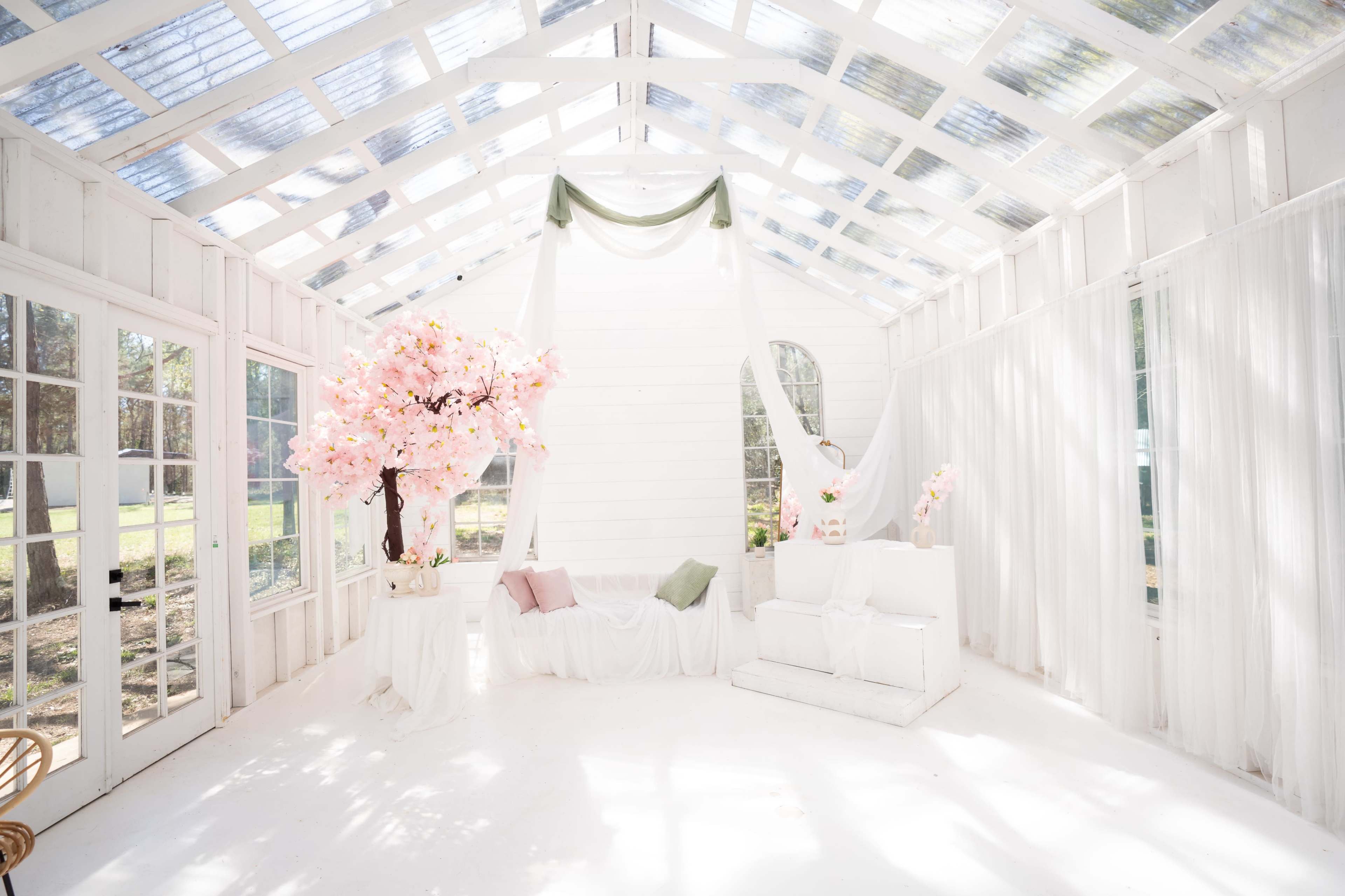 The image depicts a bright, airy white room with large windows, featuring a decorative tree with pink blossoms and minimalist furniture.
