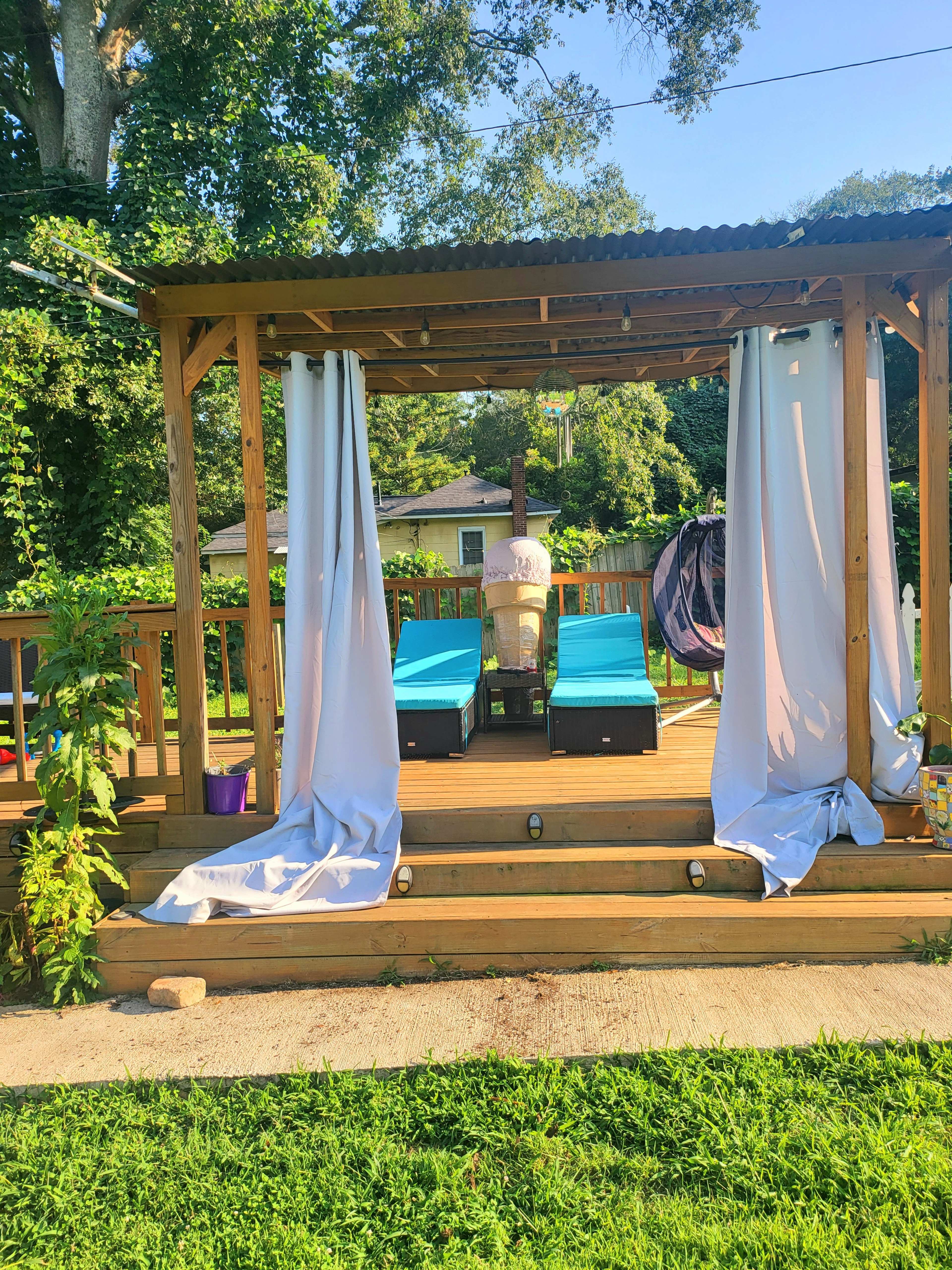 A wooden patio with two blue lounge chairs and white curtains, surrounded by greenery and a hanging seat.