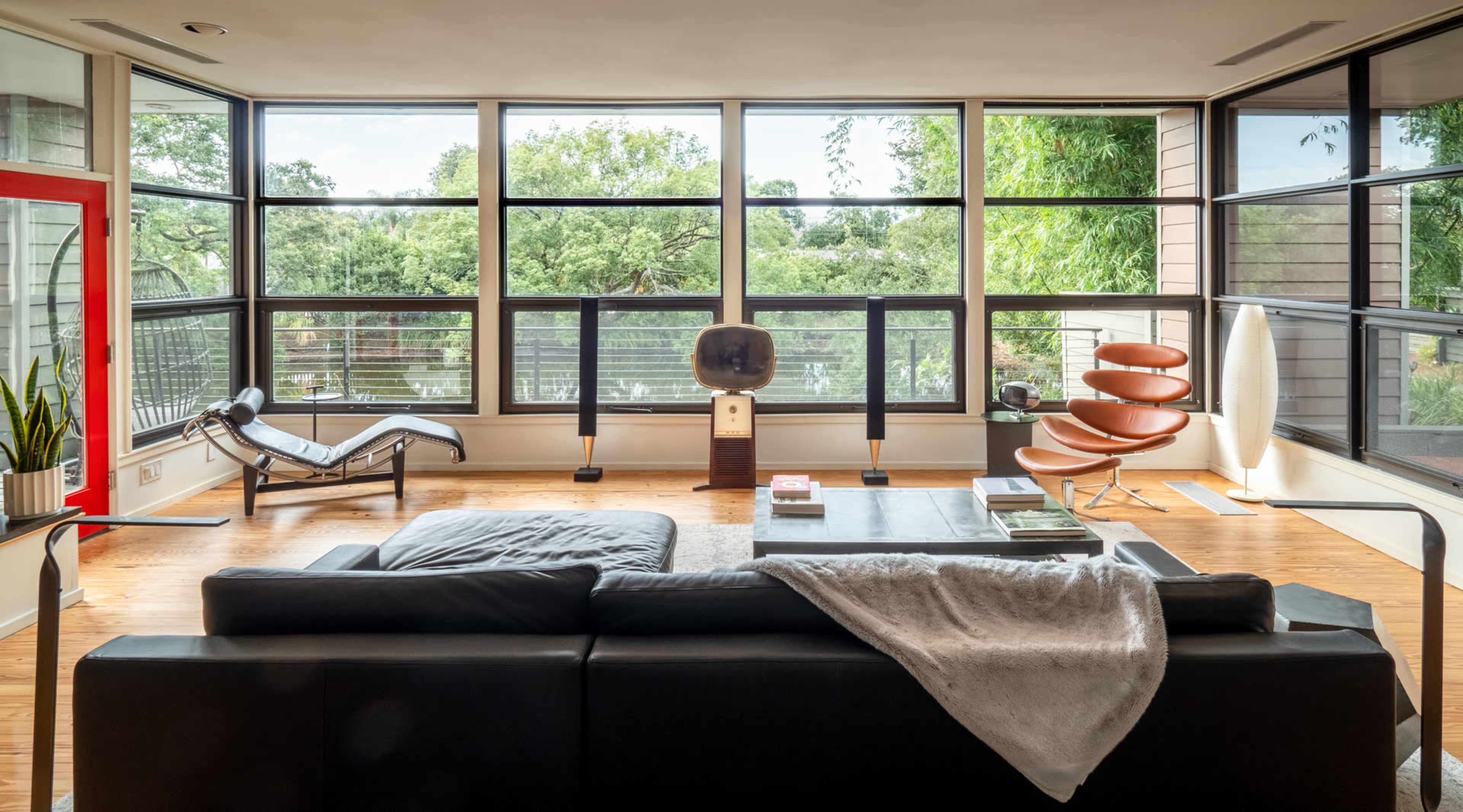 A modern living room with large windows, a black sectional sofa, and various mid-century modern furniture pieces.