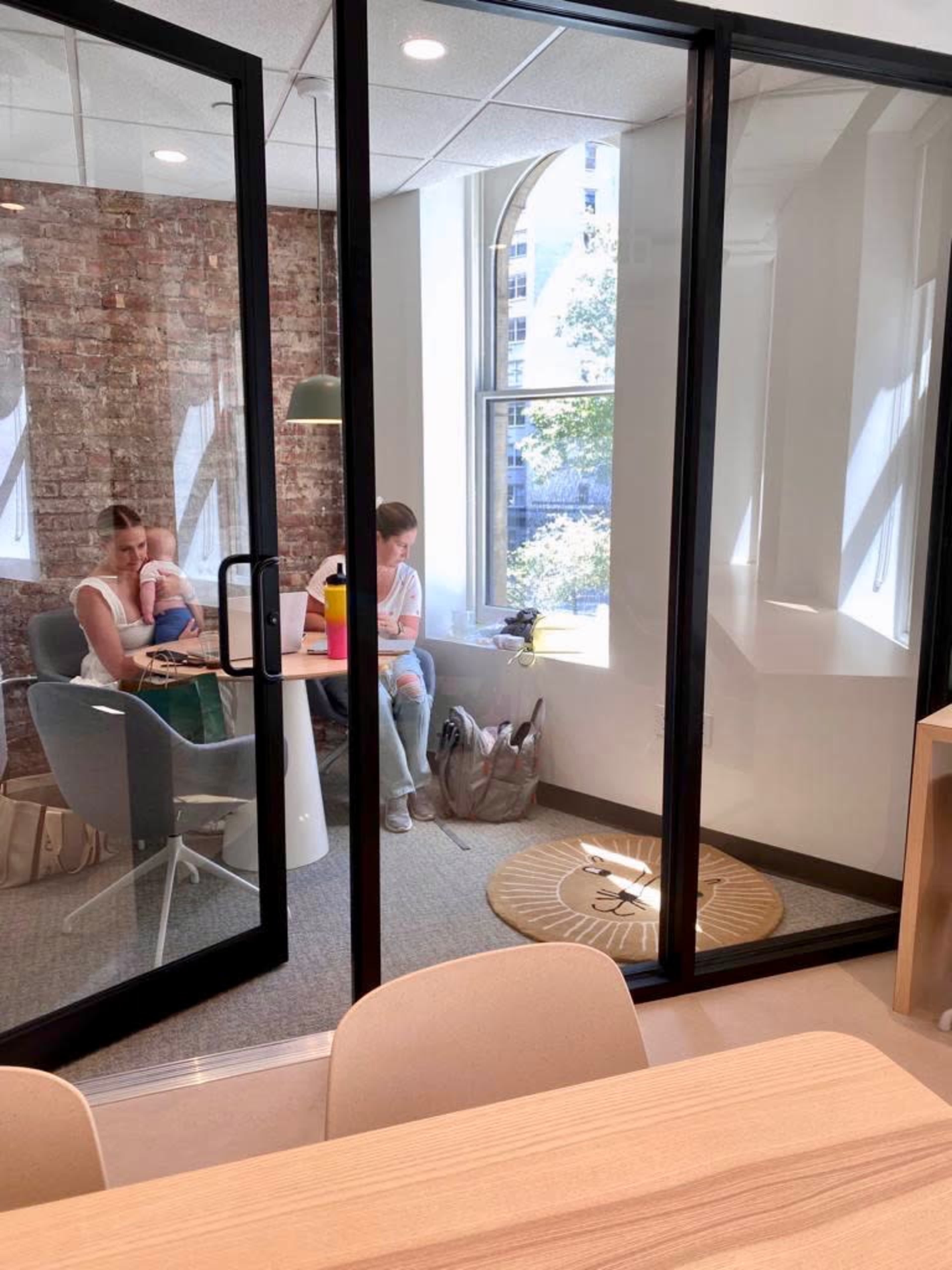 The image shows two women in a glass-walled office space, one sitting at a table and the other on the floor by a window.