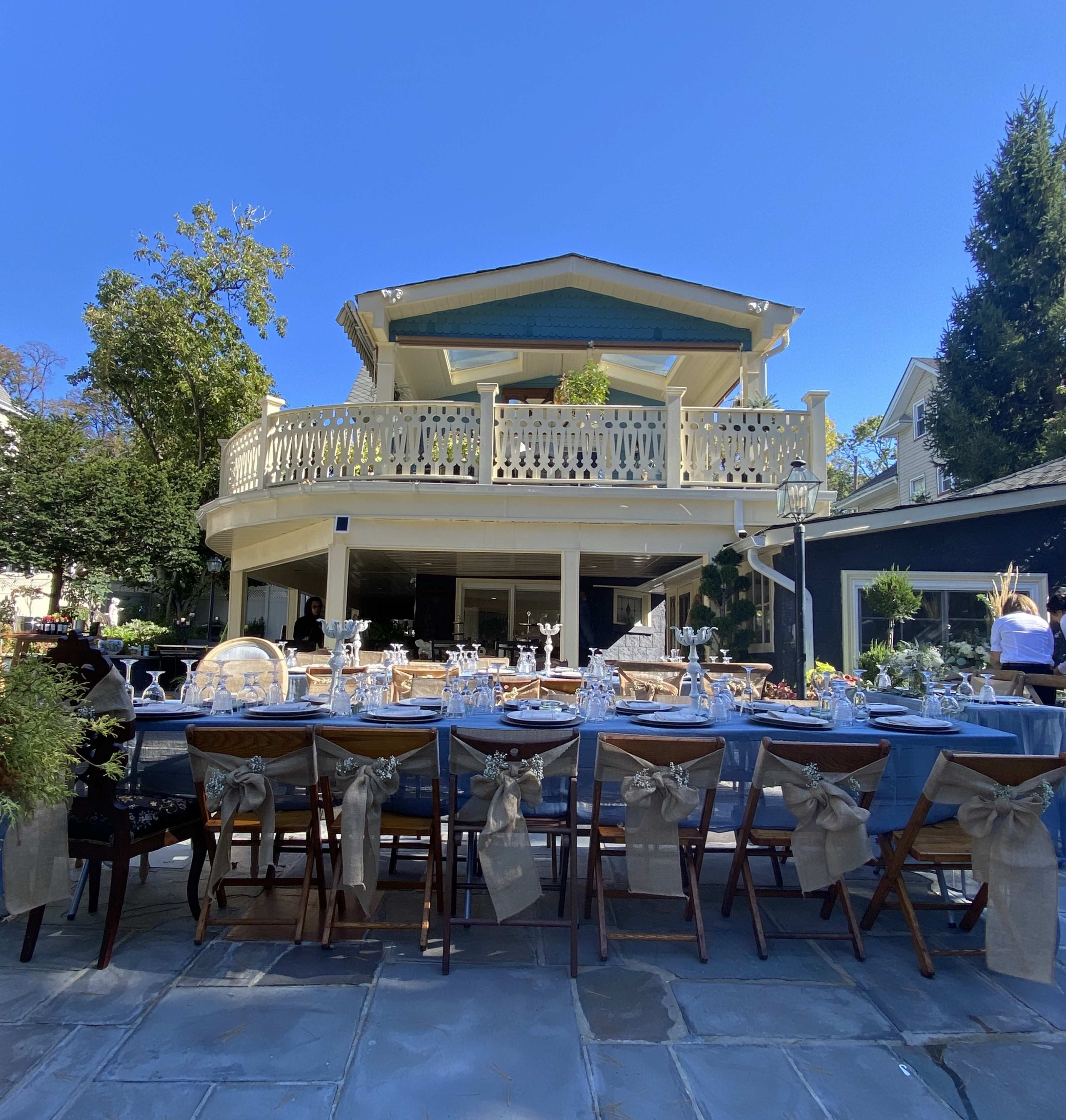 A large outdoor dining table is set with glassware and tableware in front of a multi-story house with a wraparound balcony.