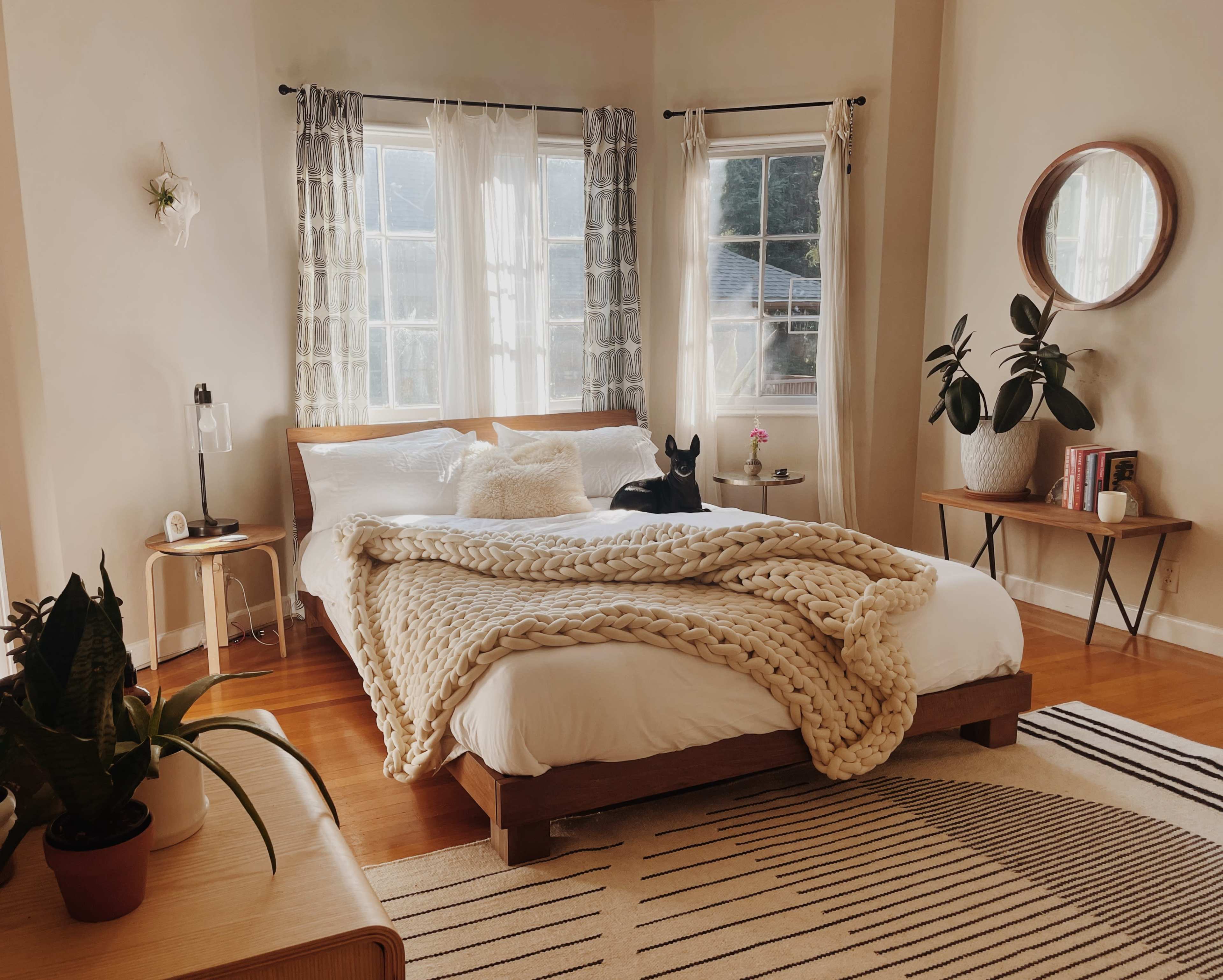 A well-lit bedroom features a large bed with a chunky knit blanket, potted plants, and two windows draped with sheer curtains.