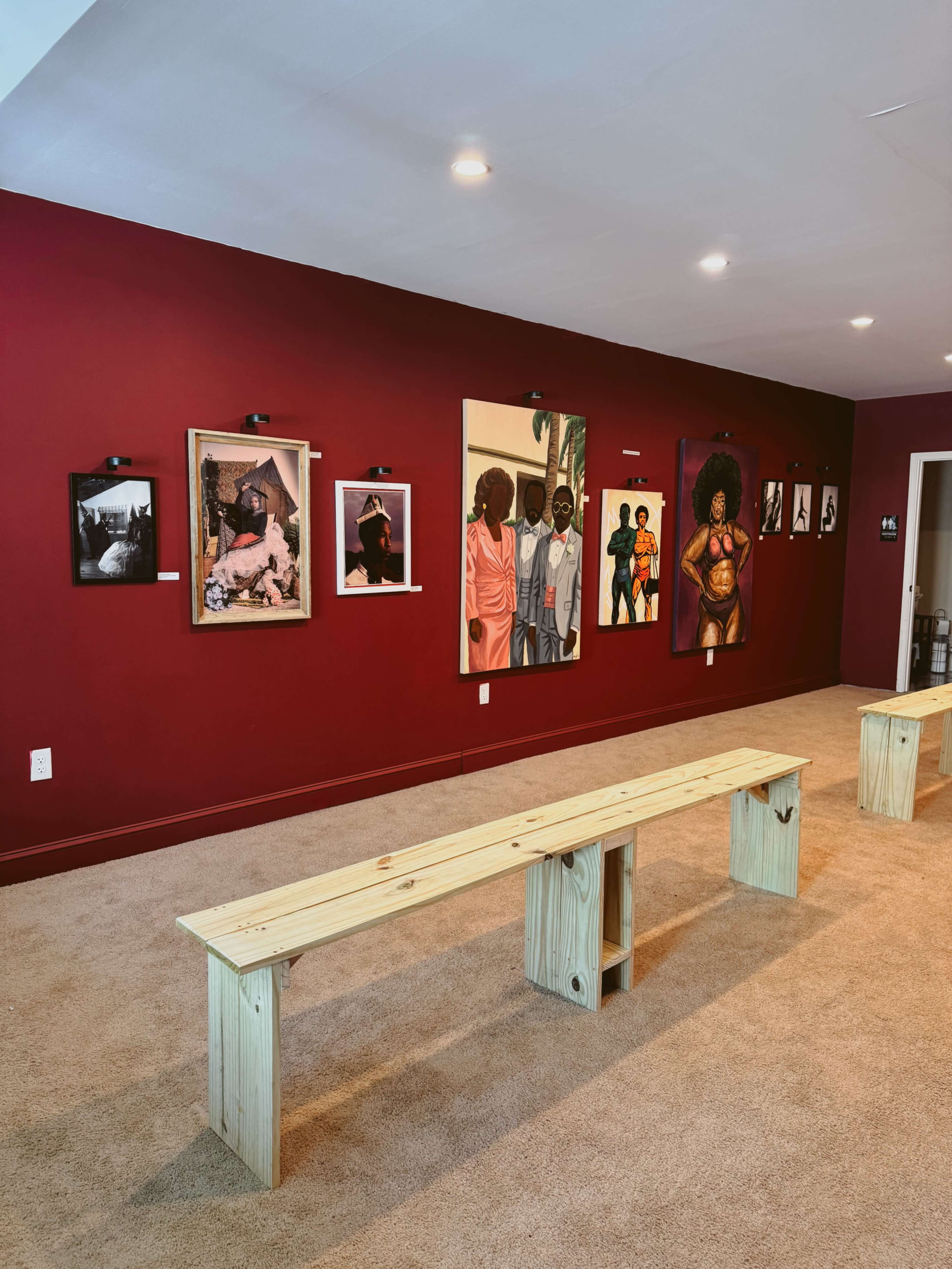 The image shows a gallery space with red walls displaying various framed artworks, alongside two wooden benches for seating.