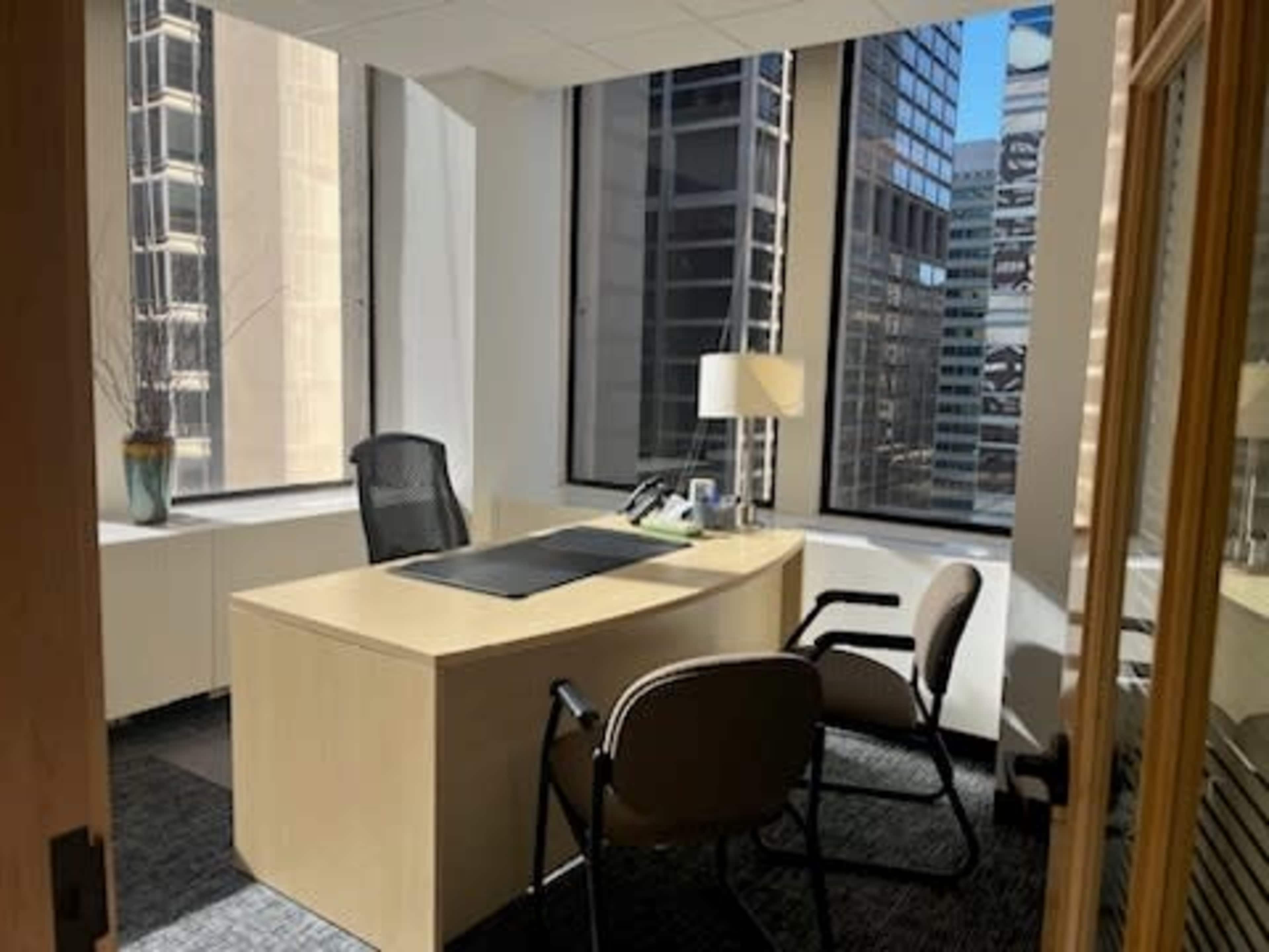 The image shows an office space with a wooden desk, two chairs, and large windows displaying a cityscape.