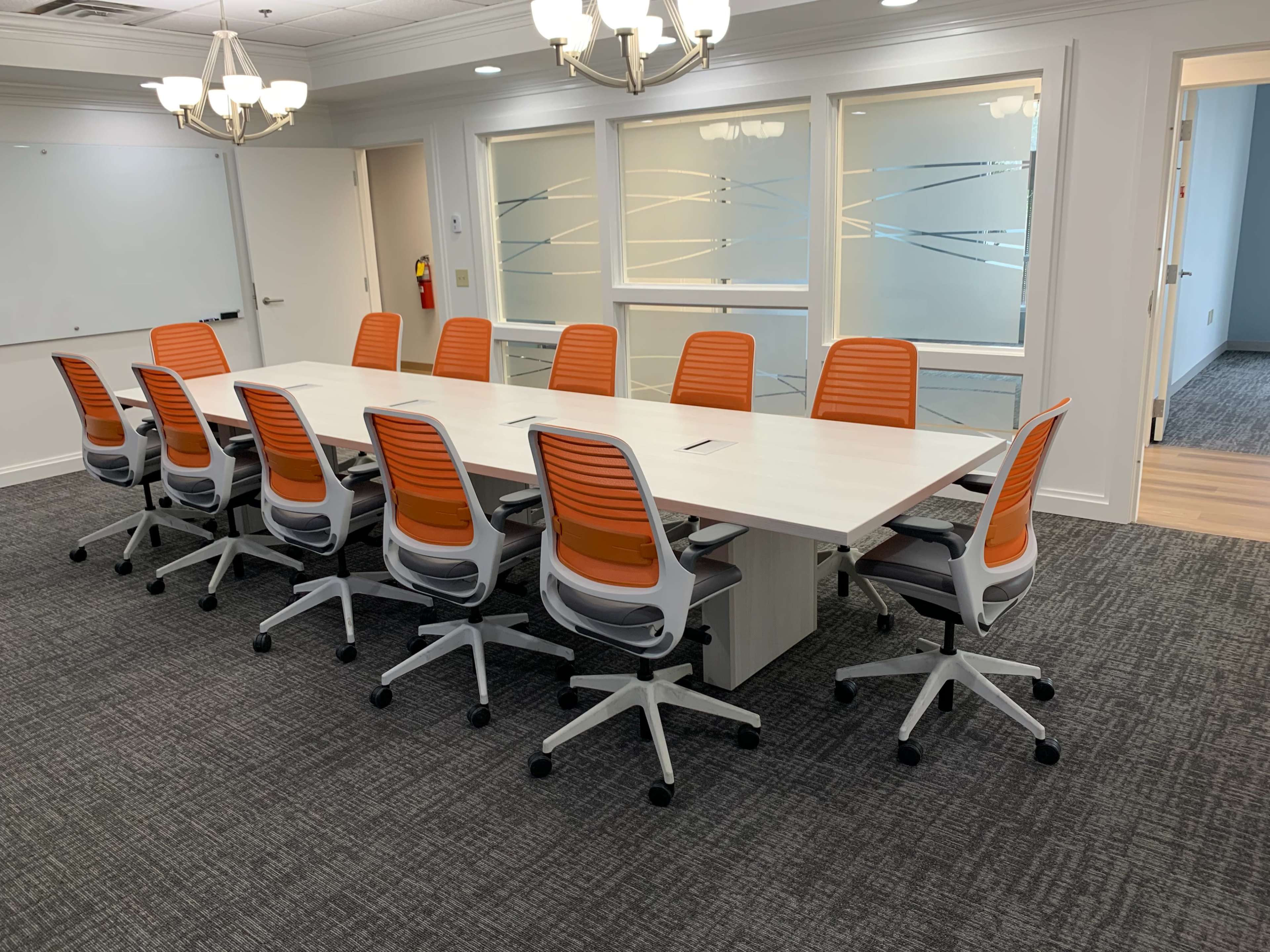 A conference room is set with a long white table surrounded by twelve orange and gray office chairs.