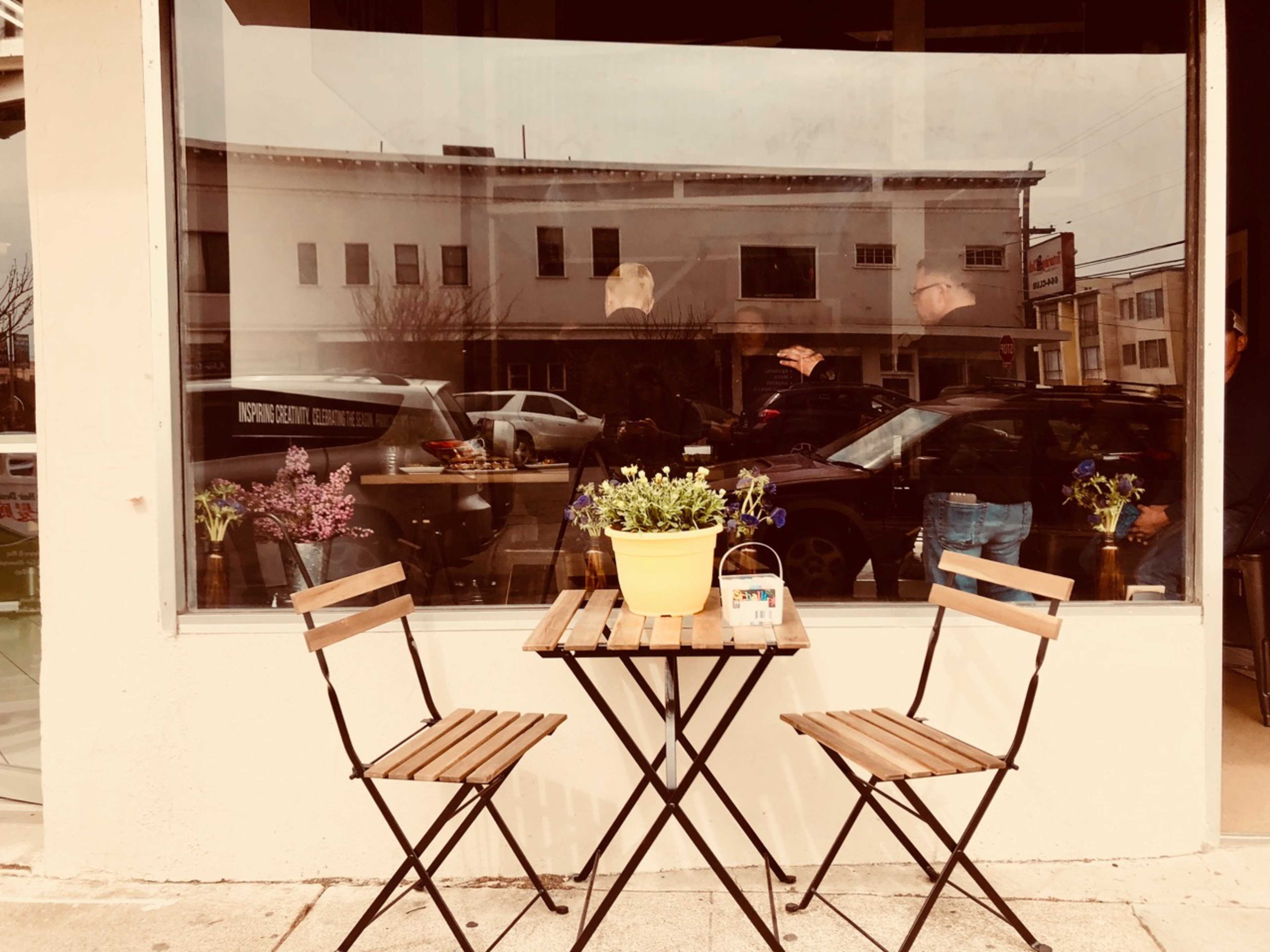A small, wooden table with two folding chairs and potted flowers is set outside a café, reflecting a building and vehicles in the window.
