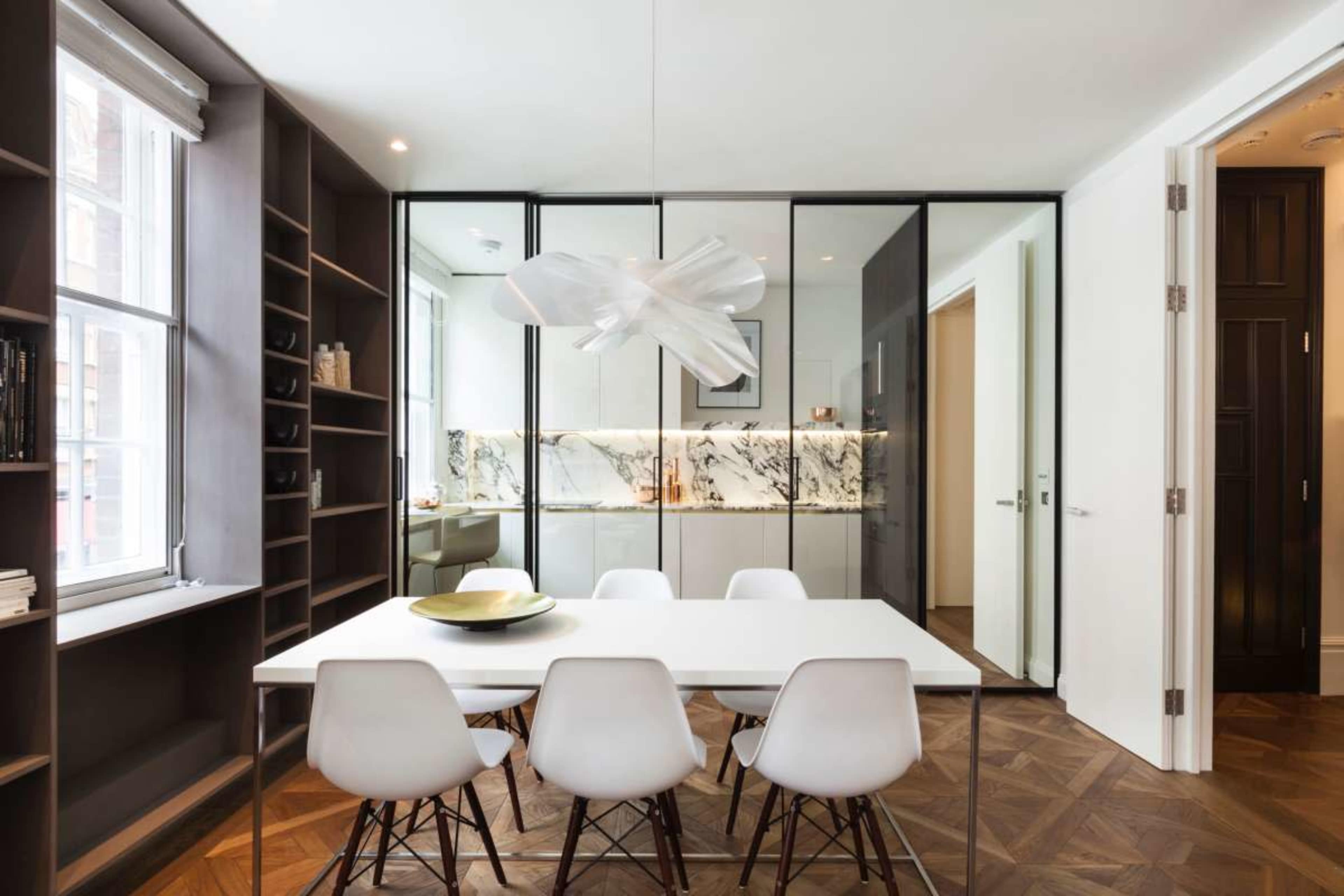 The image shows a modern dining area featuring a white table with six chairs, illuminated by a unique light fixture, along with glass dividers and a marble backsplash in the background.