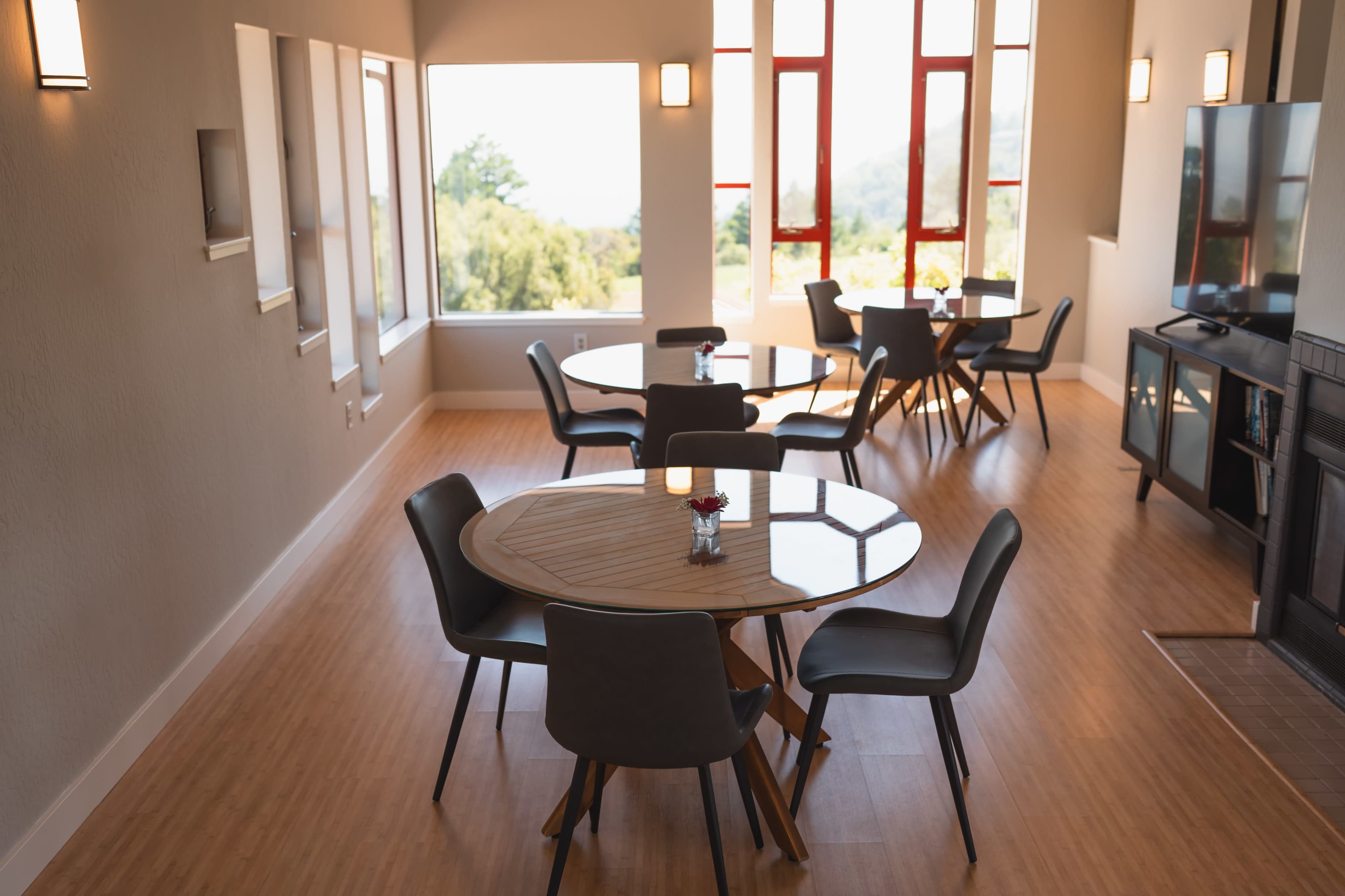 The image shows a spacious room with several round tables surrounded by chairs, large windows allowing natural light, and a view of greenery outside.