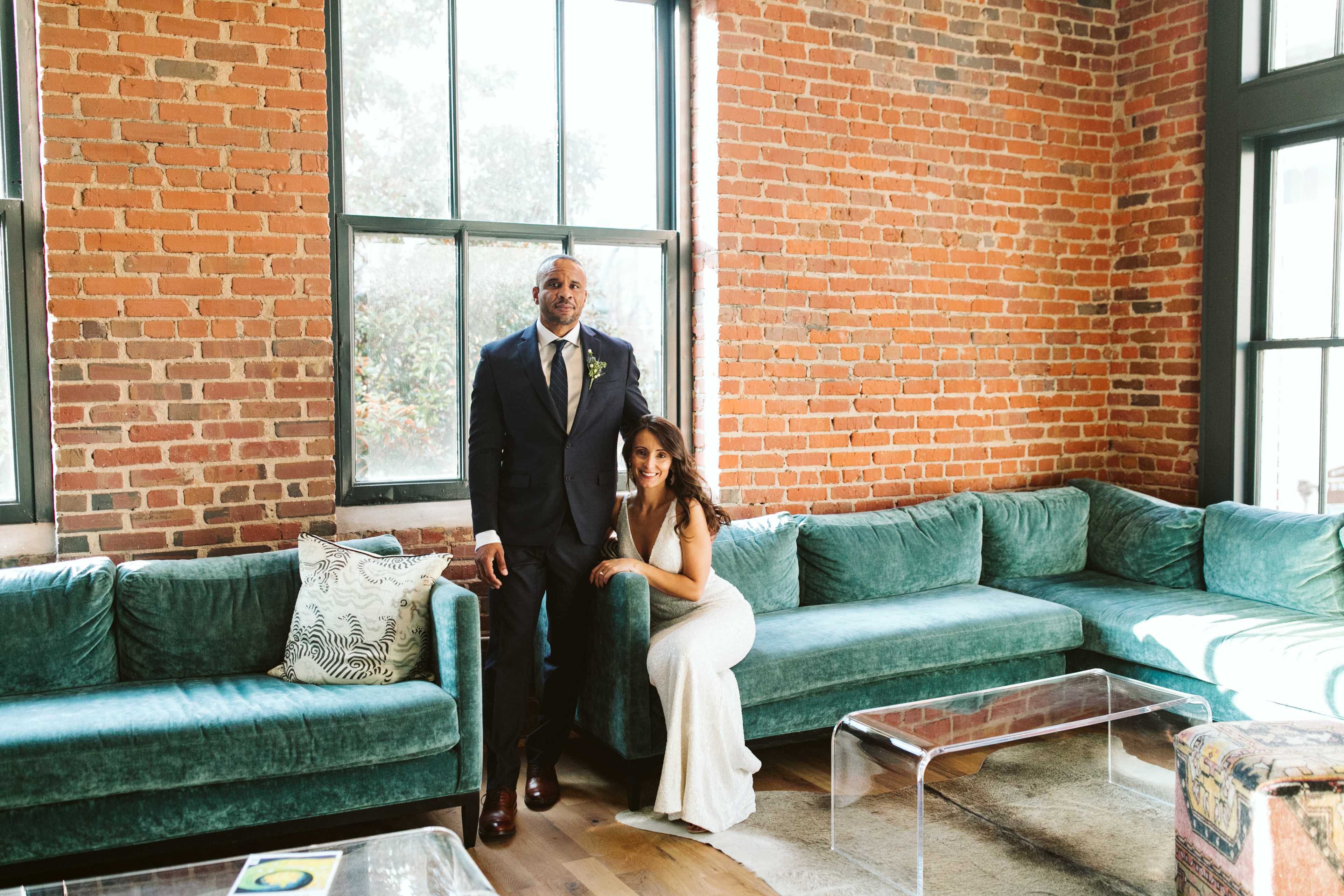 A man in a suit stands beside a woman in a white dress in a room with exposed brick walls and teal sofas.