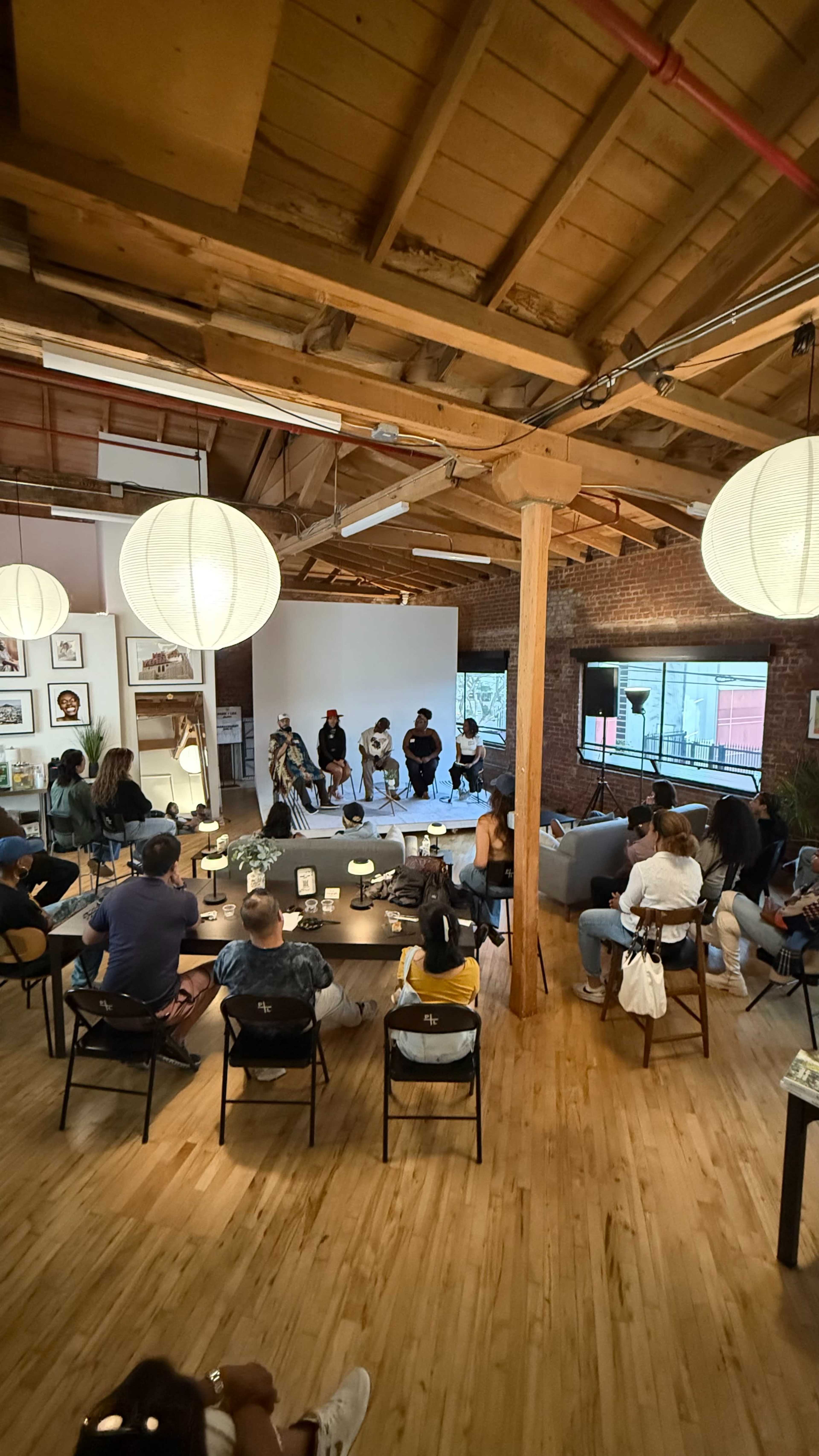 A group of people is seated in a spacious, well-lit venue for a panel discussion, with a small stage at the front and large paper lanterns hanging from the ceiling.