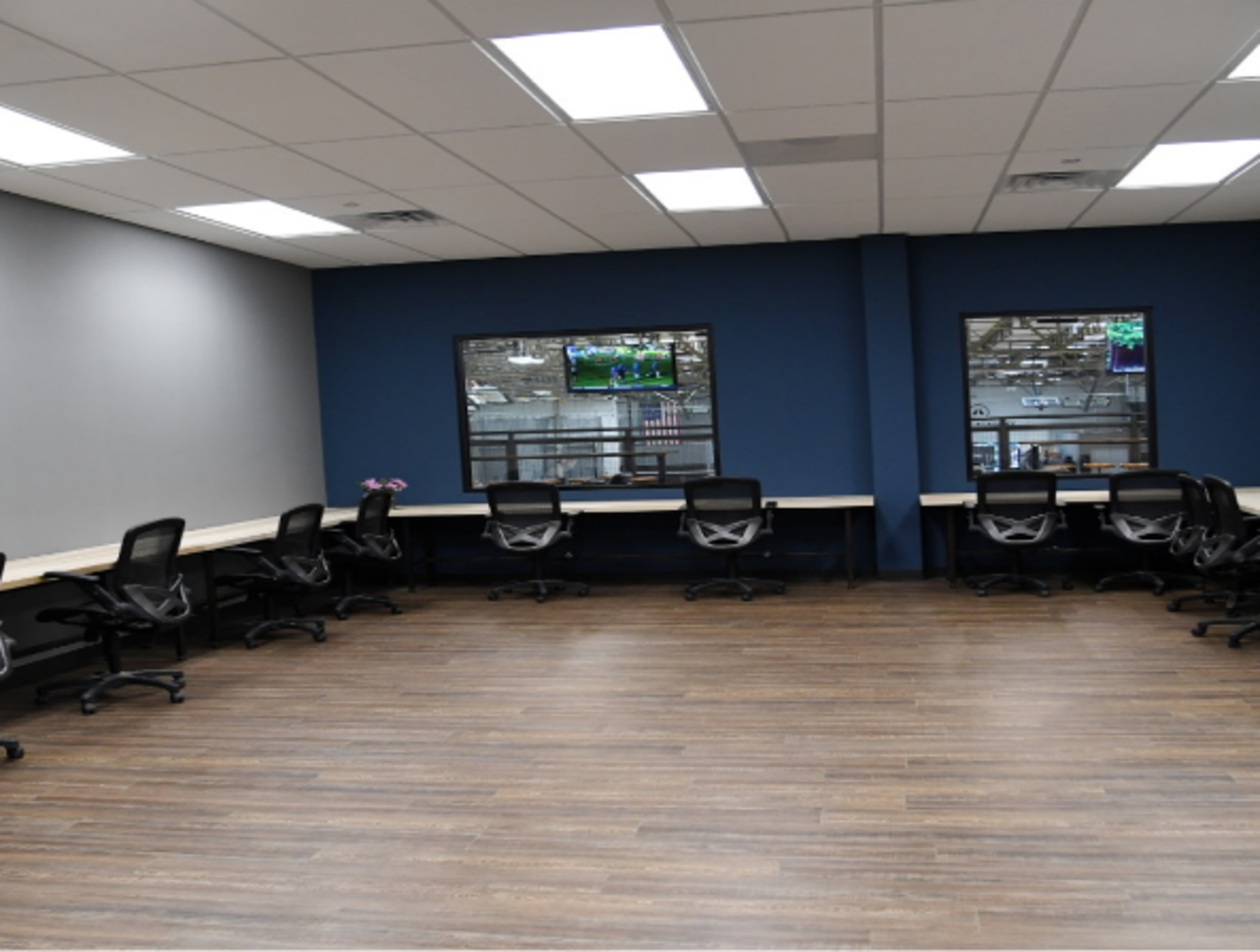 The image shows a modern office space with several ergonomic chairs facing a long table along the walls, and large windows displaying a view of a sports event.