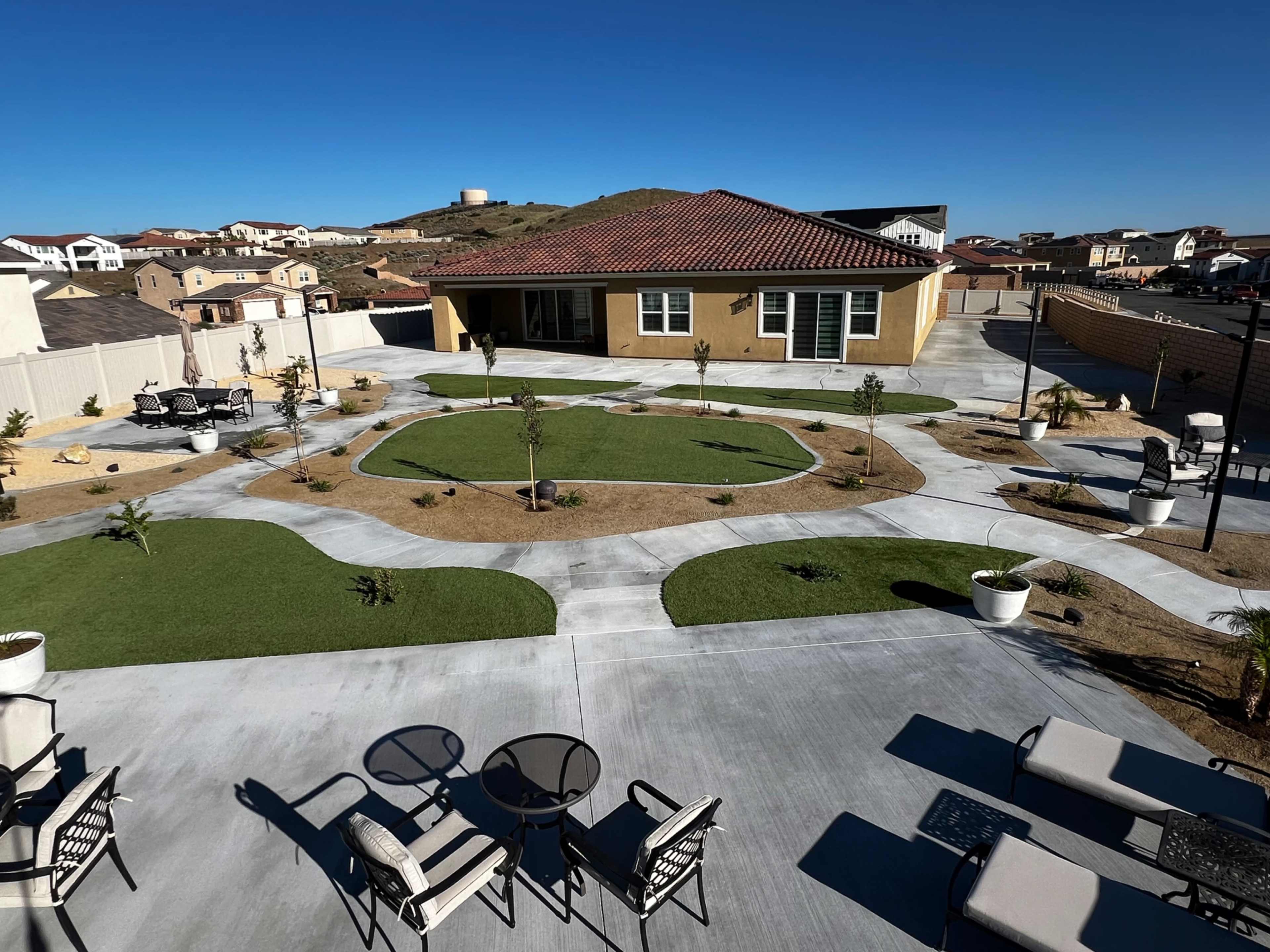 The image shows a landscaped outdoor area featuring a modern building, concrete pathways, and neatly arranged patches of grass and shrubs.