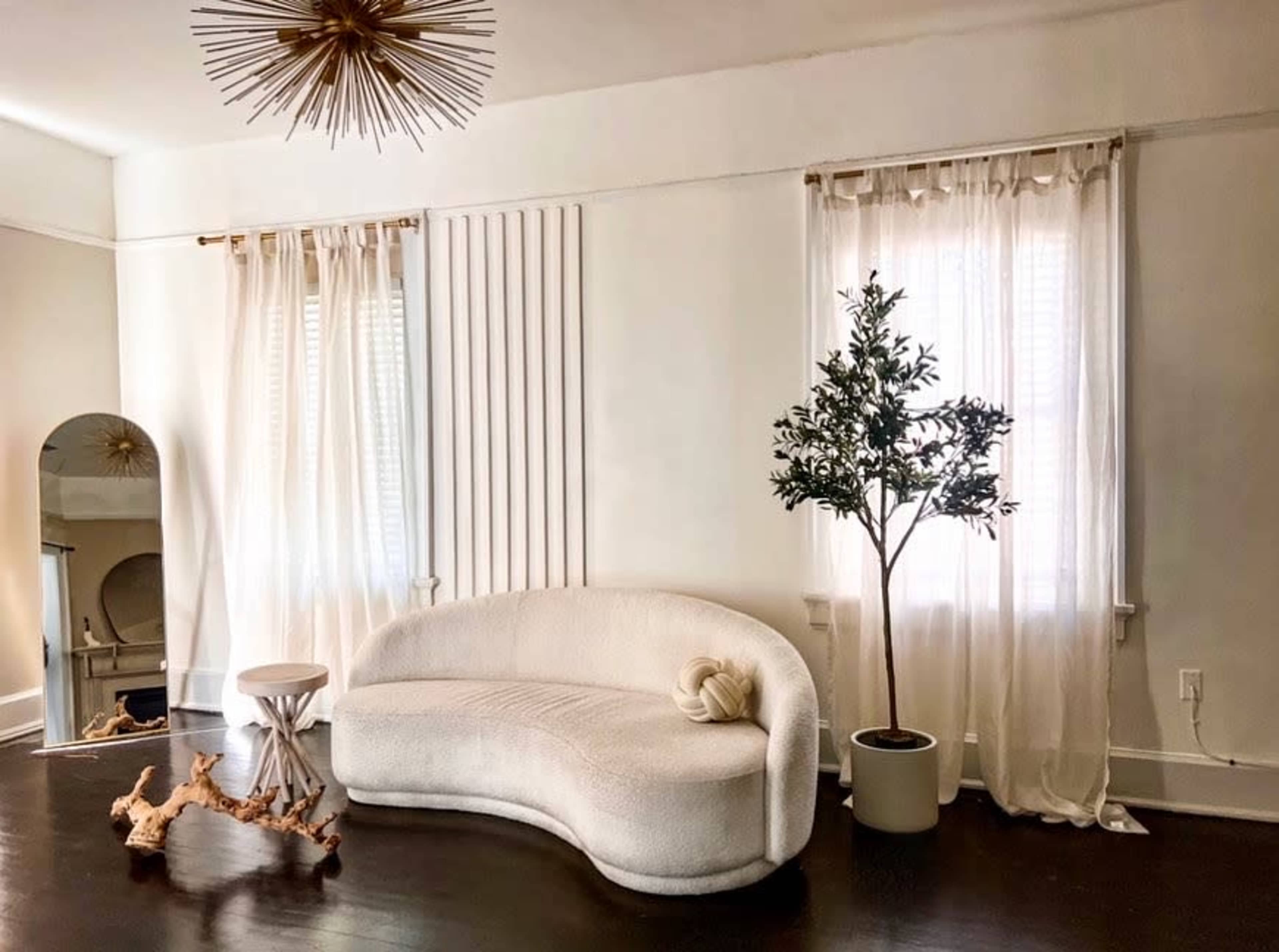 A modern living room features a curved white sofa, a potted plant, a small table, and sheer curtains covering the windows.