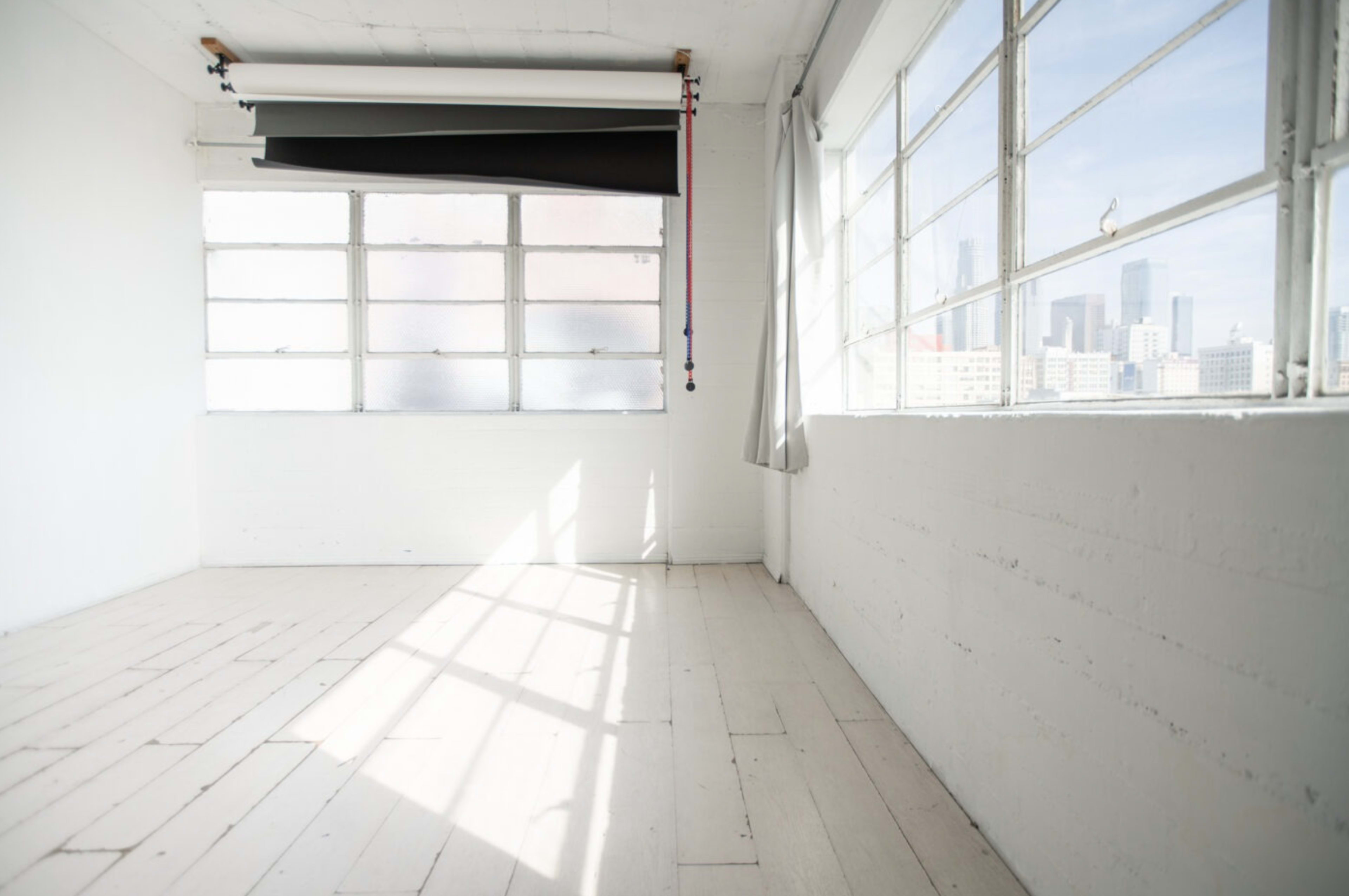 The image shows a bright, empty room with large windows and white walls, featuring a roll-up black curtain near the ceiling.