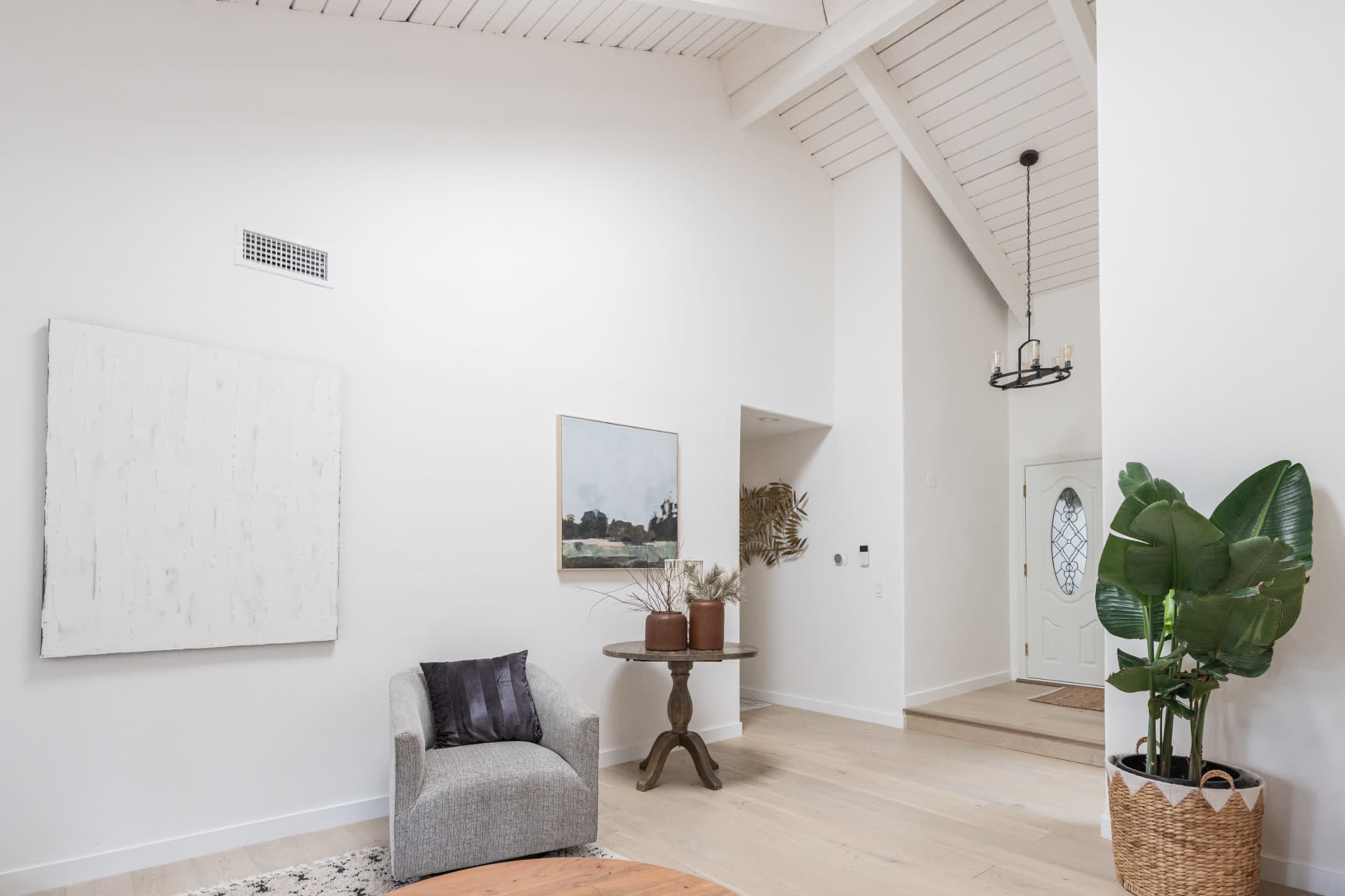 A bright, spacious living area with a gray armchair, a round table, and a large piece of artwork on the wall, complemented by a doorway leading to an entry hall.