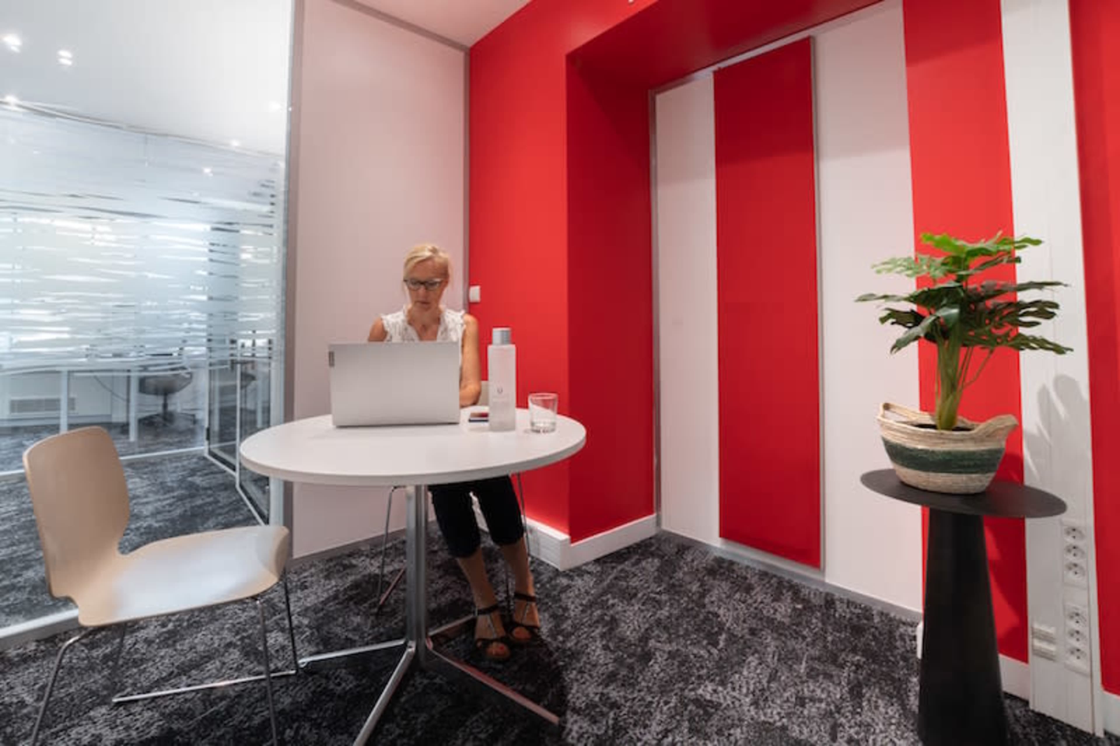 A woman is sitting at a round table with a laptop in a modern office space featuring red and white walls and a potted plant nearby.