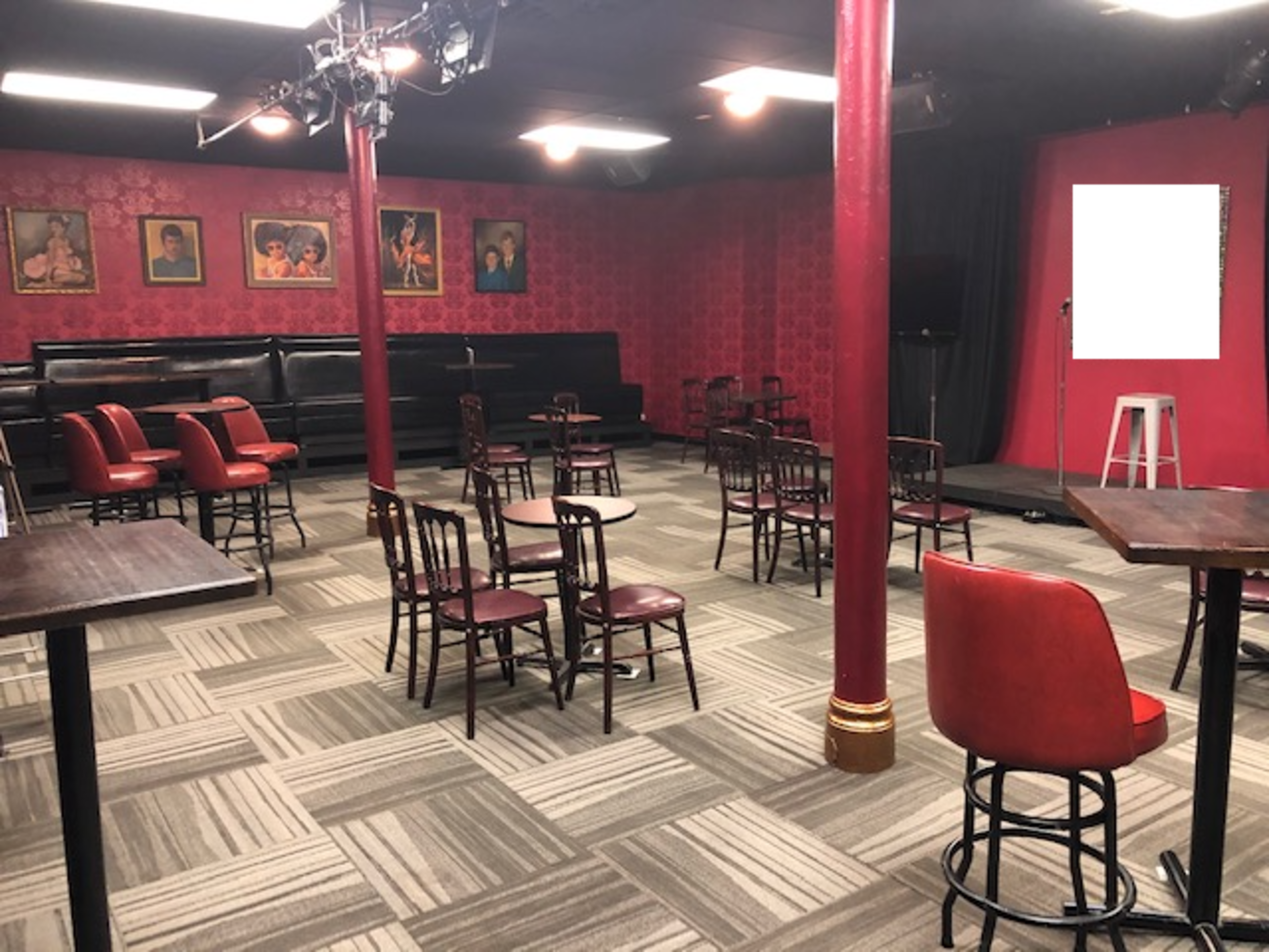 A dimly lit comedy club interior with red walls, empty tables and chairs, and a small stage at the back.
