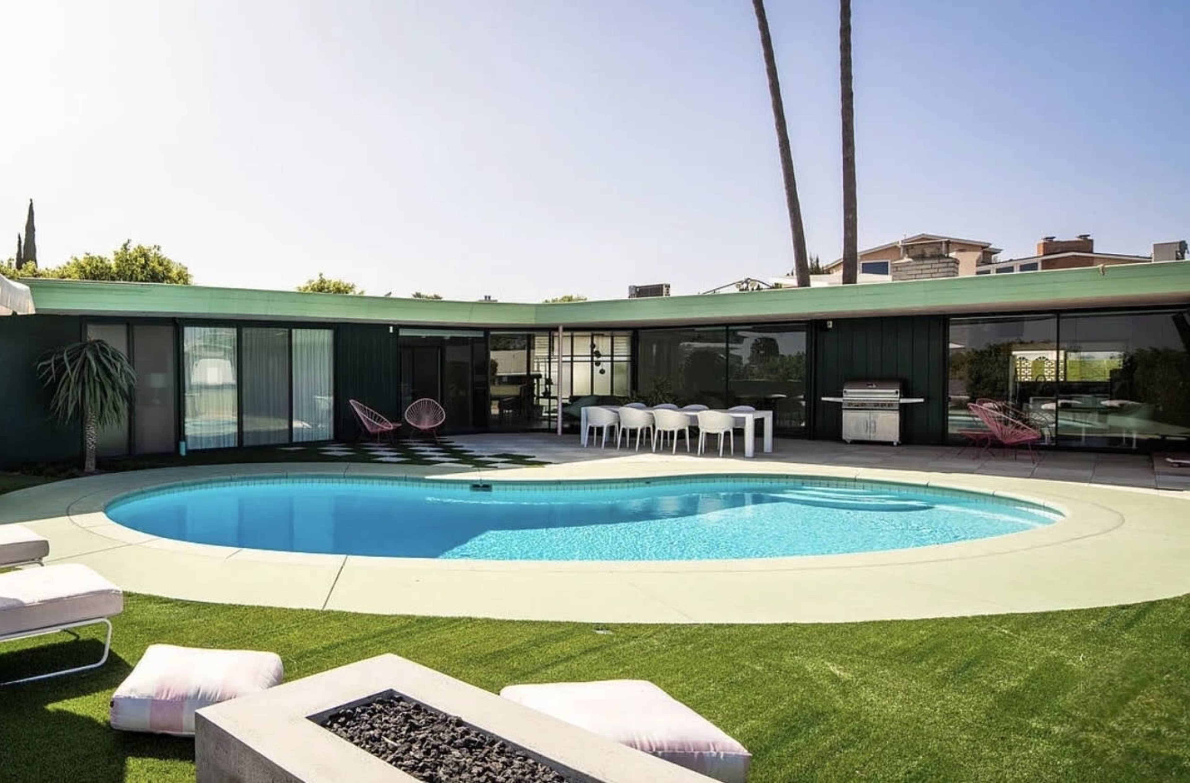 The image shows a modern, circular swimming pool surrounded by a green lawn, lounge chairs, and a spacious patio area with outdoor furniture and a barbecue.