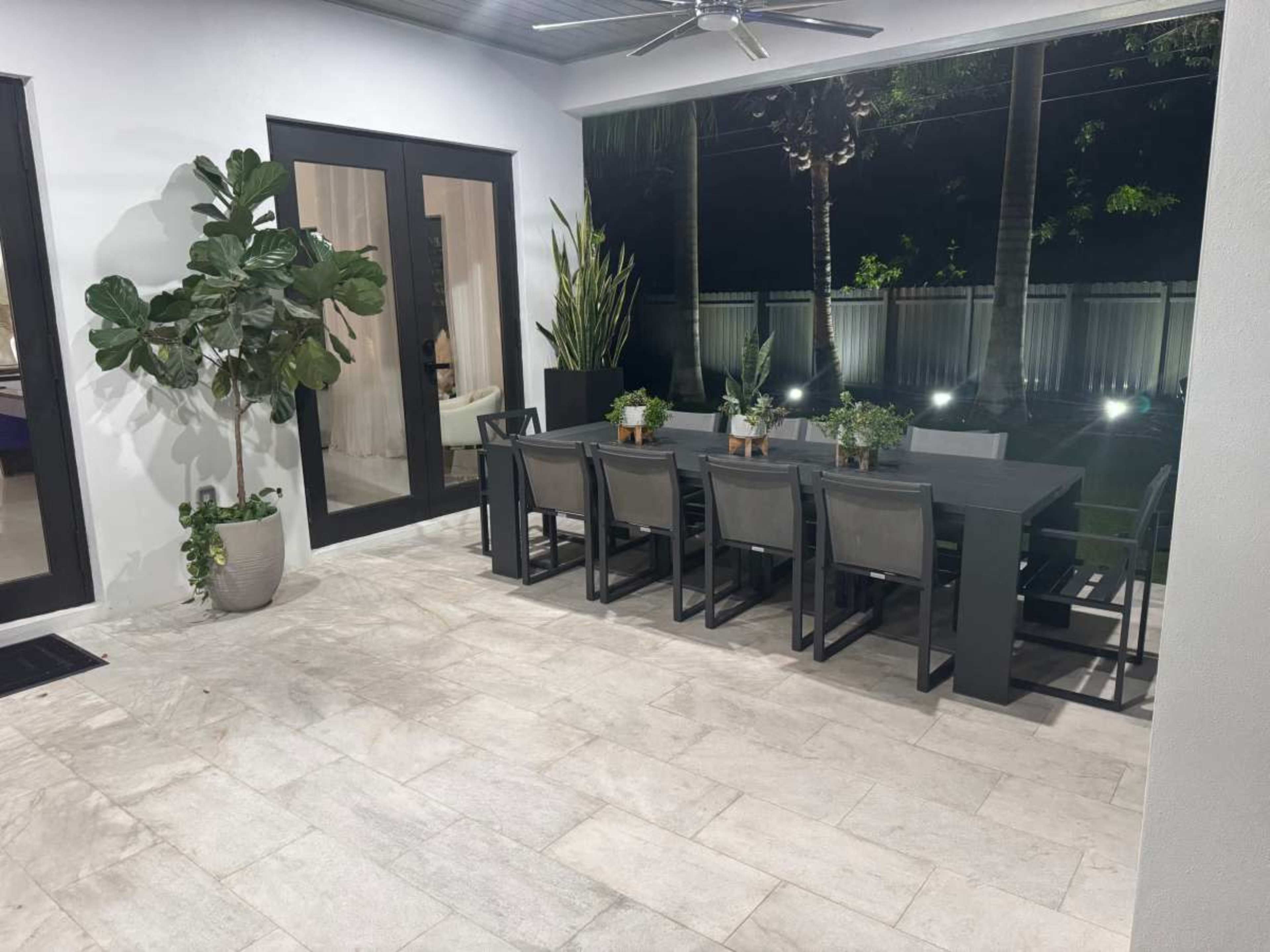 A spacious outdoor dining area features a long table with chairs, surrounded by potted plants and illuminated by soft lighting during the evening.