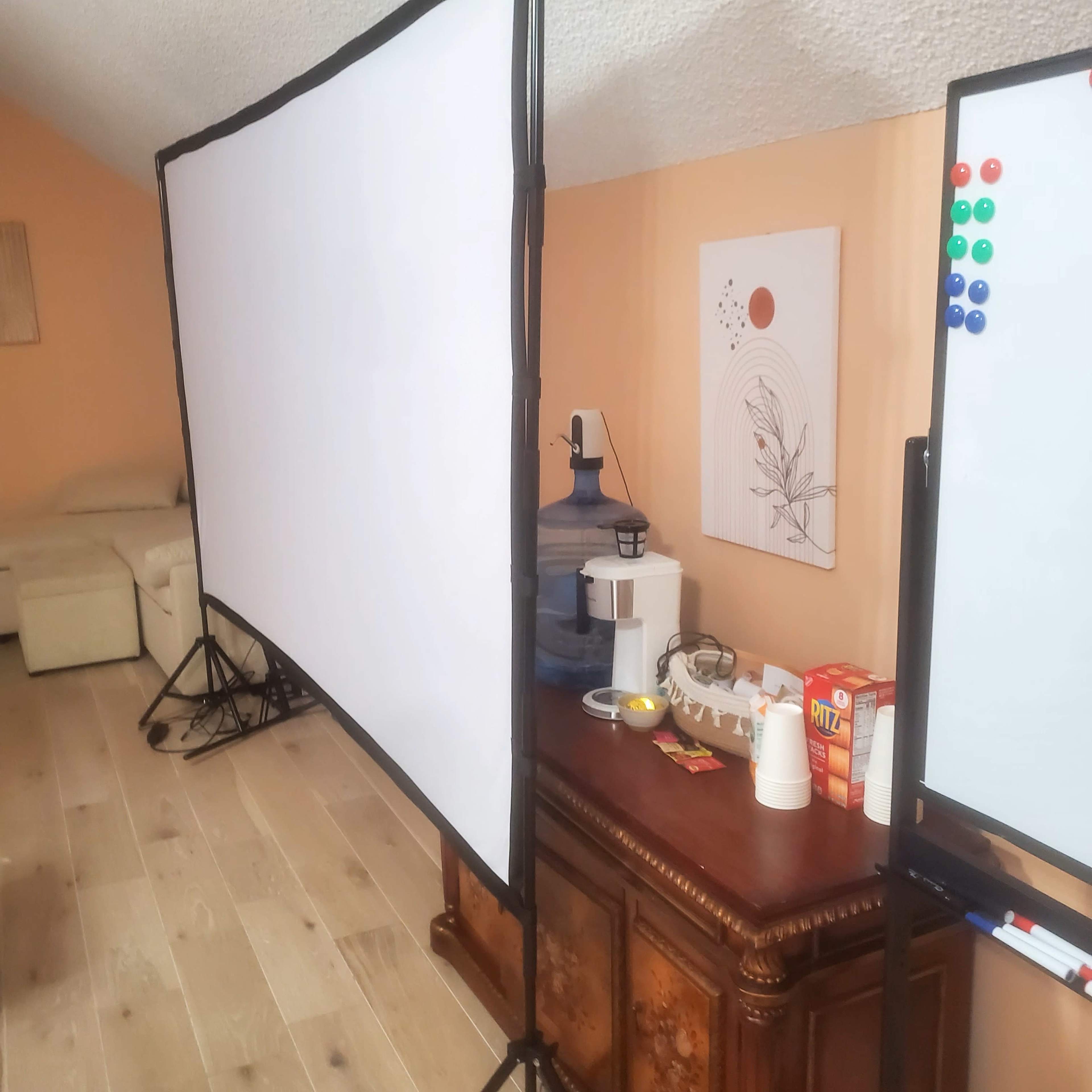 A room with two large projection screens set up, a water cooler, a whiteboard, and snacks on a wooden table.