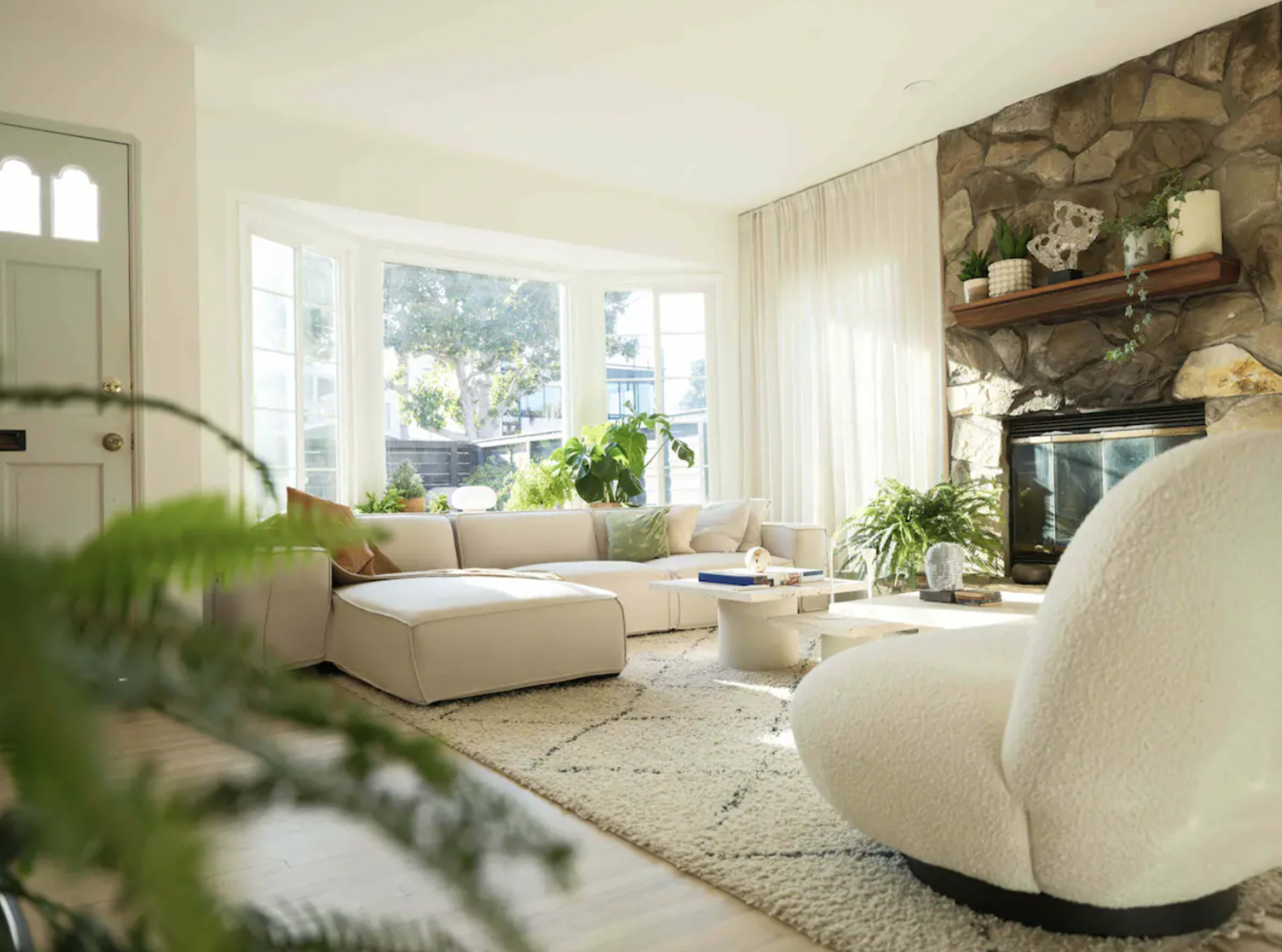 A modern living room features a light sofa, a stone fireplace, large windows, and various plants.