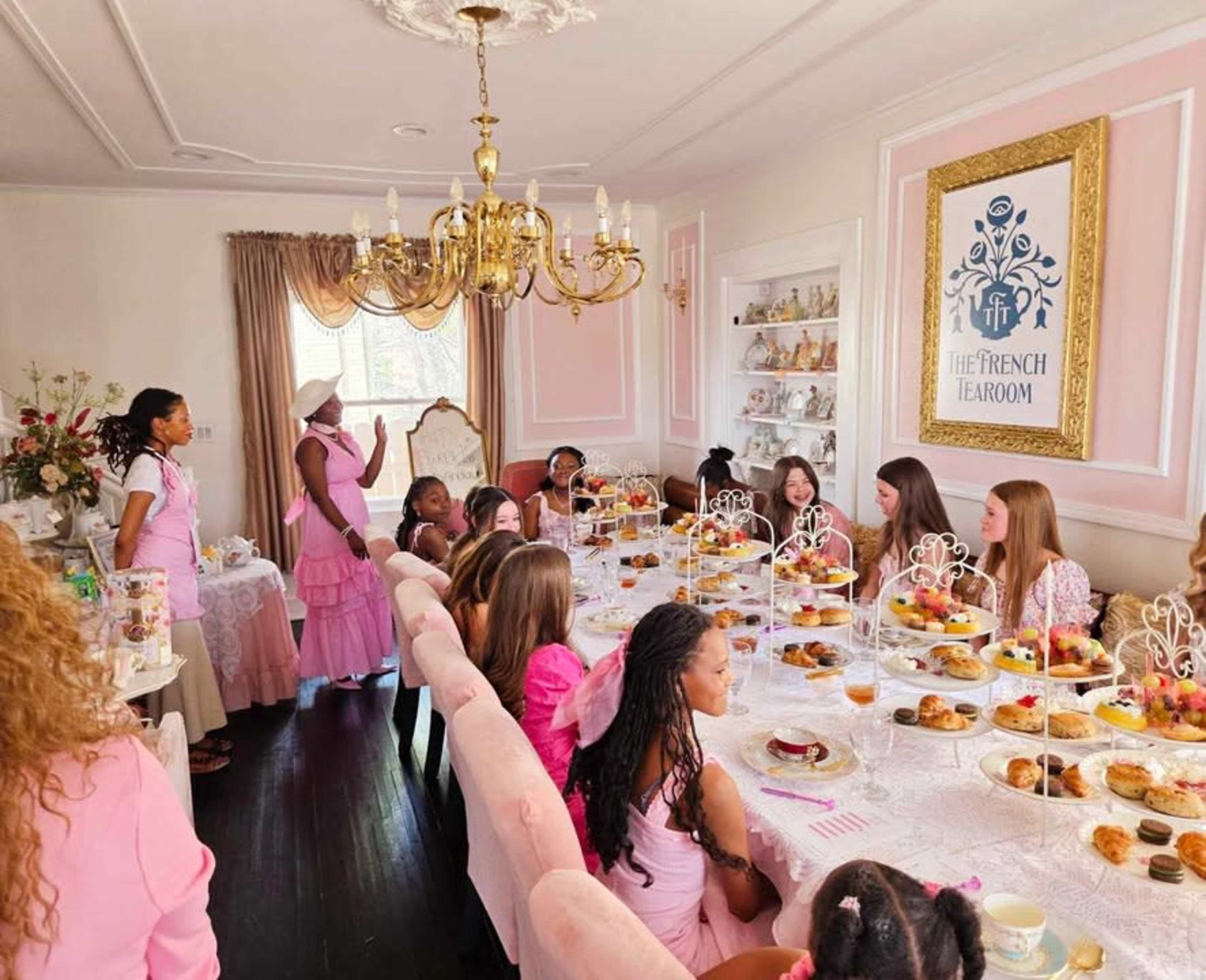 A group of young girls in pink attire is gathered around a table filled with pastries and tea in a elegantly decorated room.