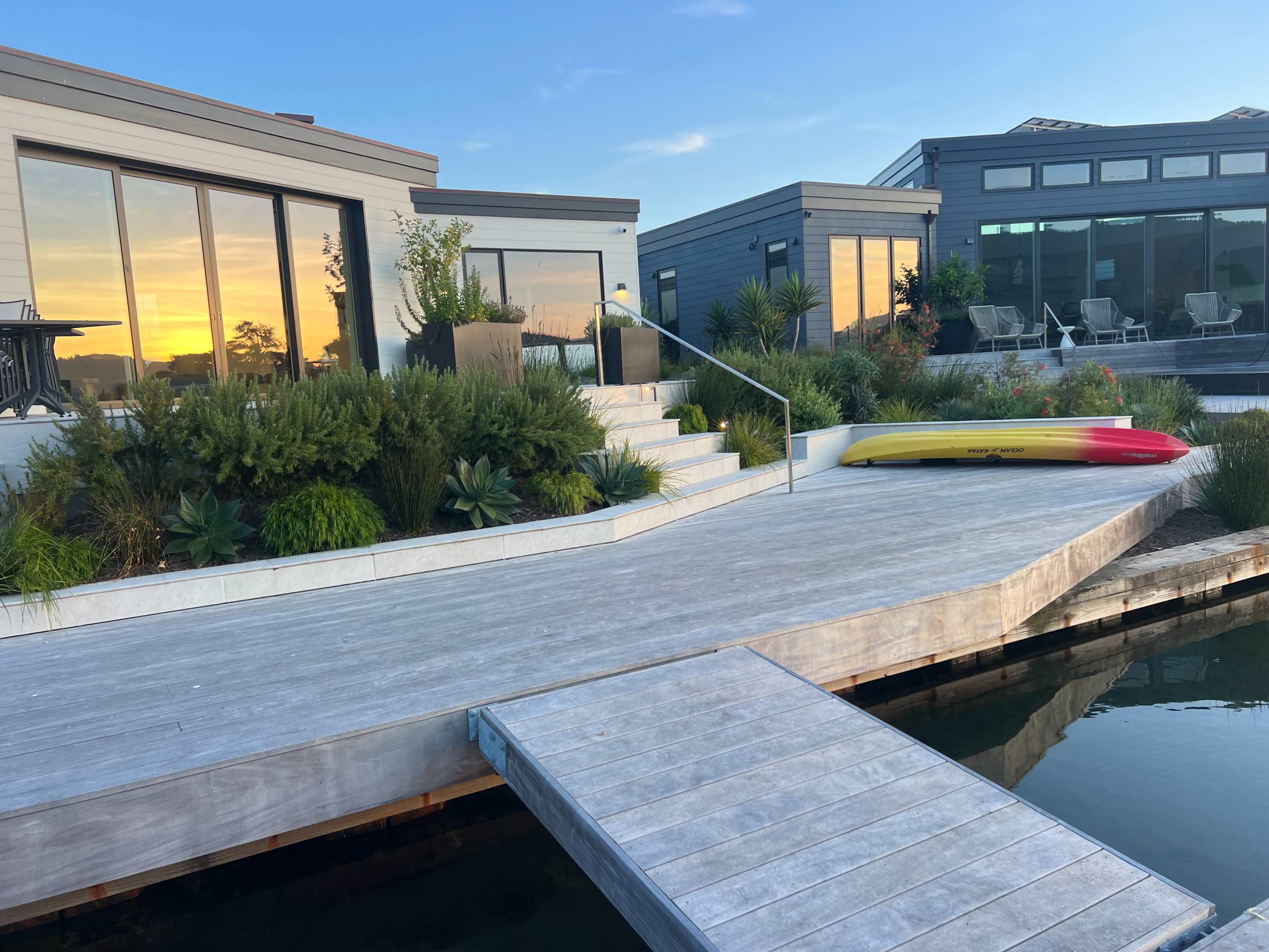 The image shows a modern waterfront property with a wooden deck extending into the water, featuring lush greenery, and a kayak resting on the side.