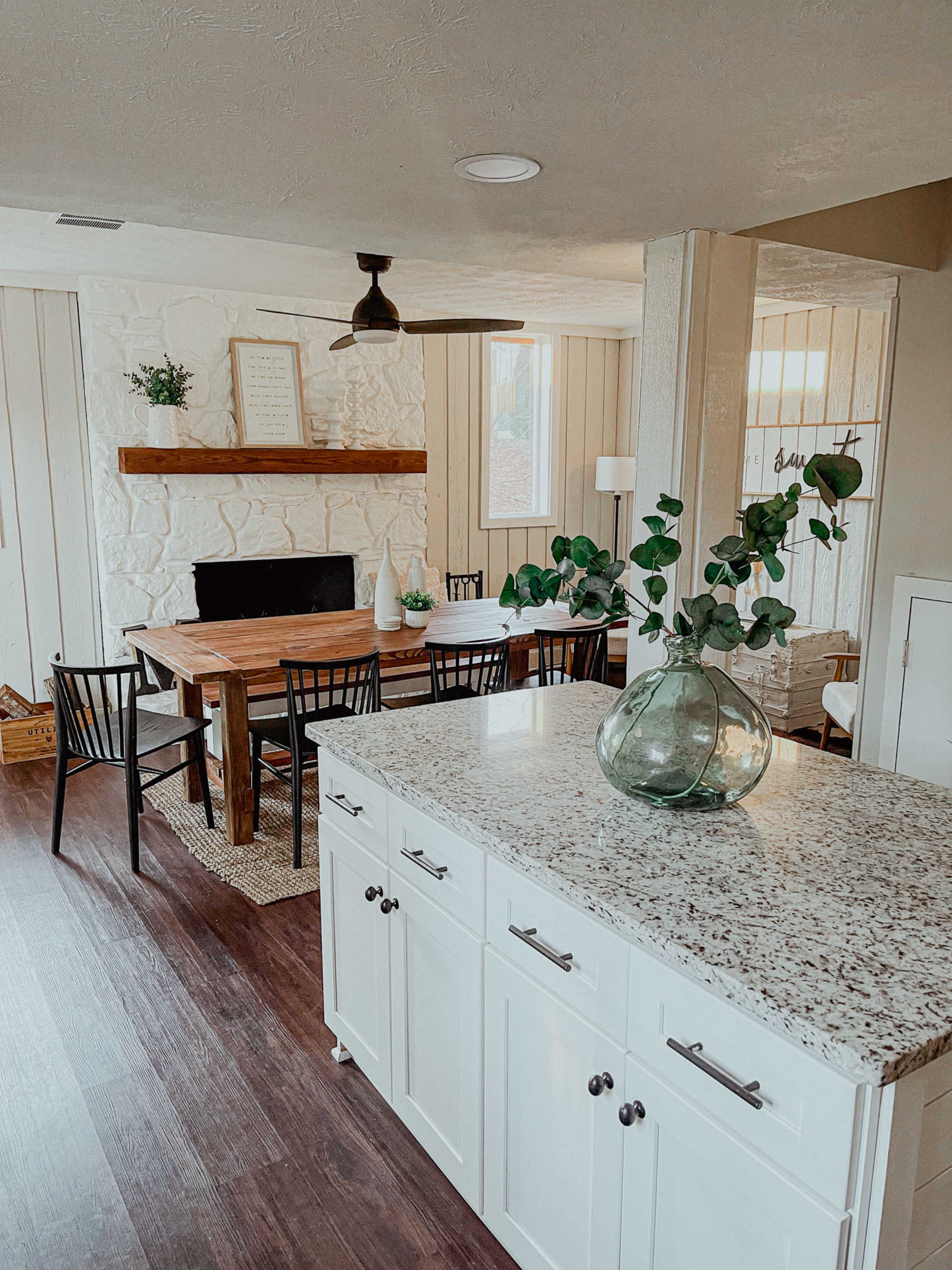 The image shows a modern dining area featuring a wooden table, black chairs, a stone fireplace with a wooden mantel, and a kitchen island with a granite countertop.
