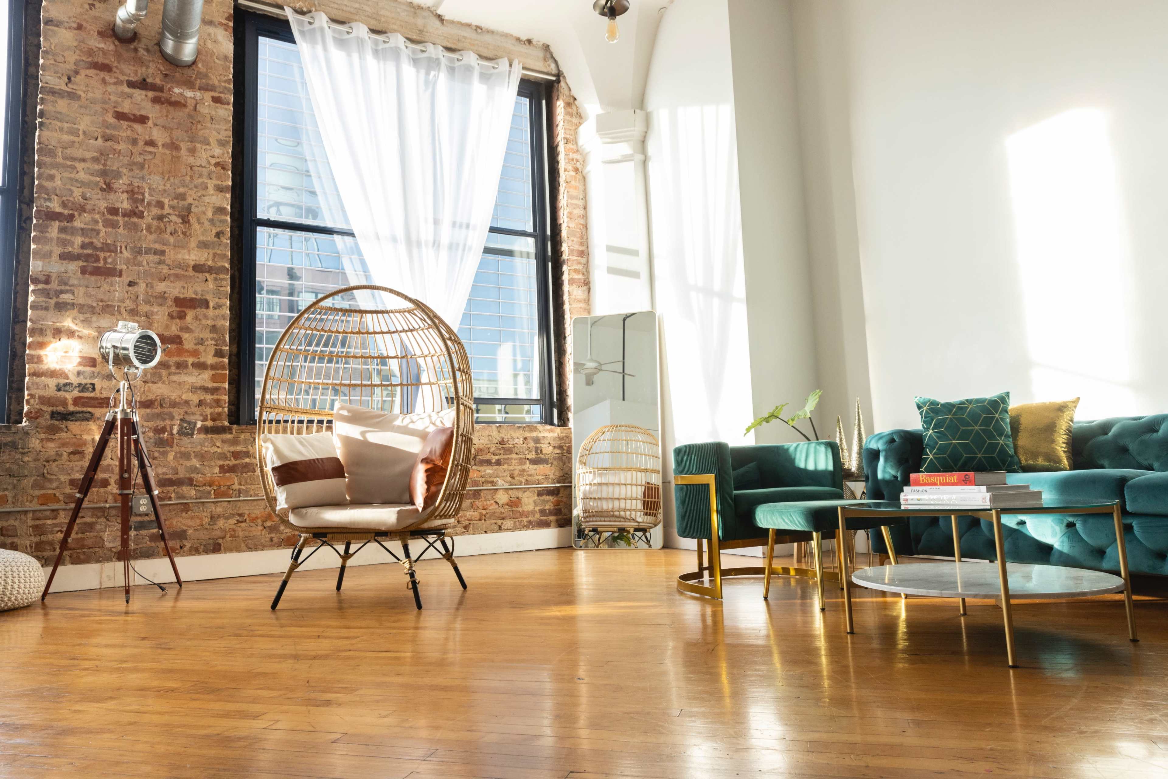 A bright, airy room featuring a rattan hanging chair, a teal sofa, and a coffee table on polished wooden flooring, with large windows letting in natural light.