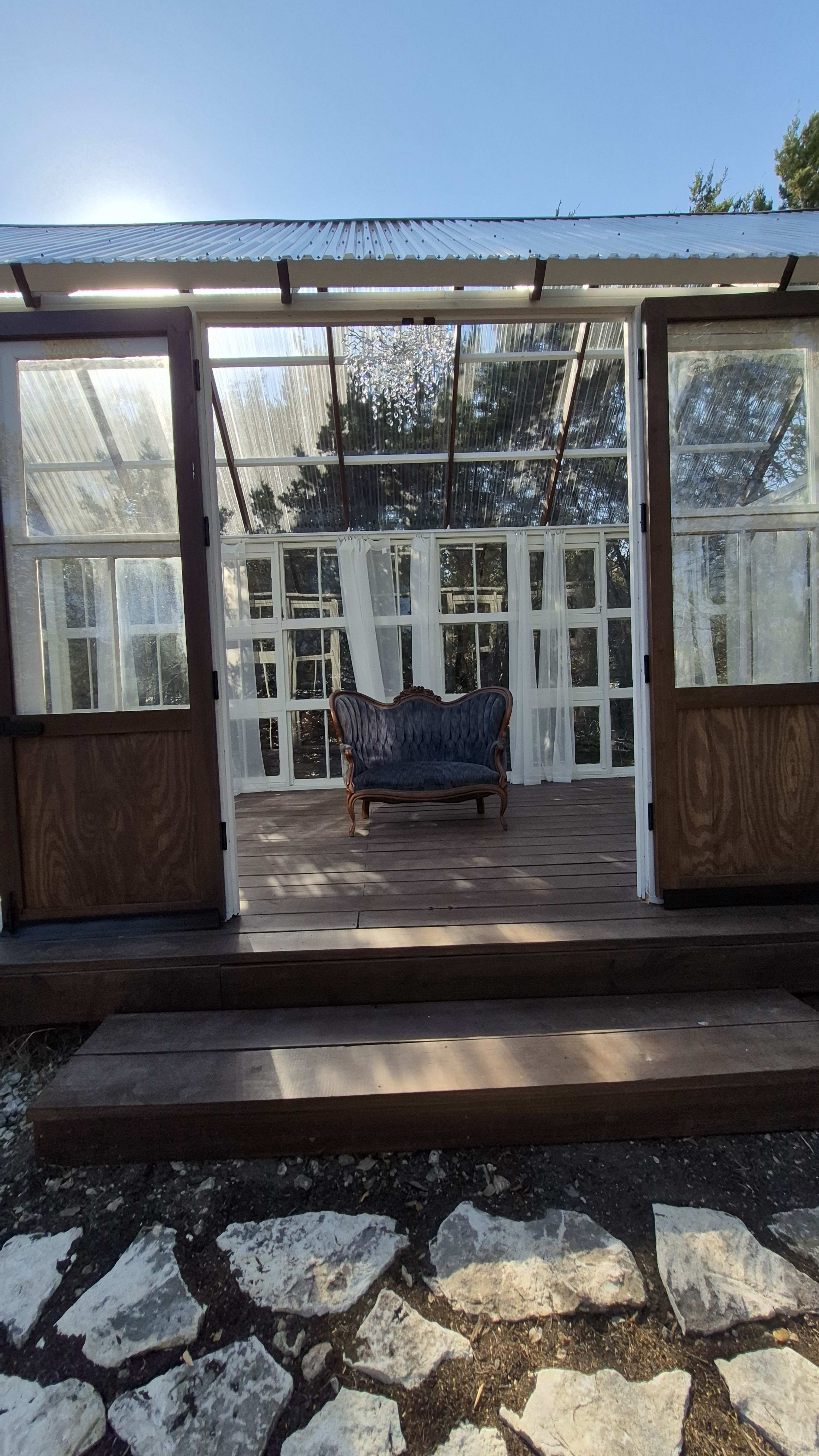 A wooden structure with large windows and a translucent roof features a vintage couch placed in the center, surrounded by open space and natural light.