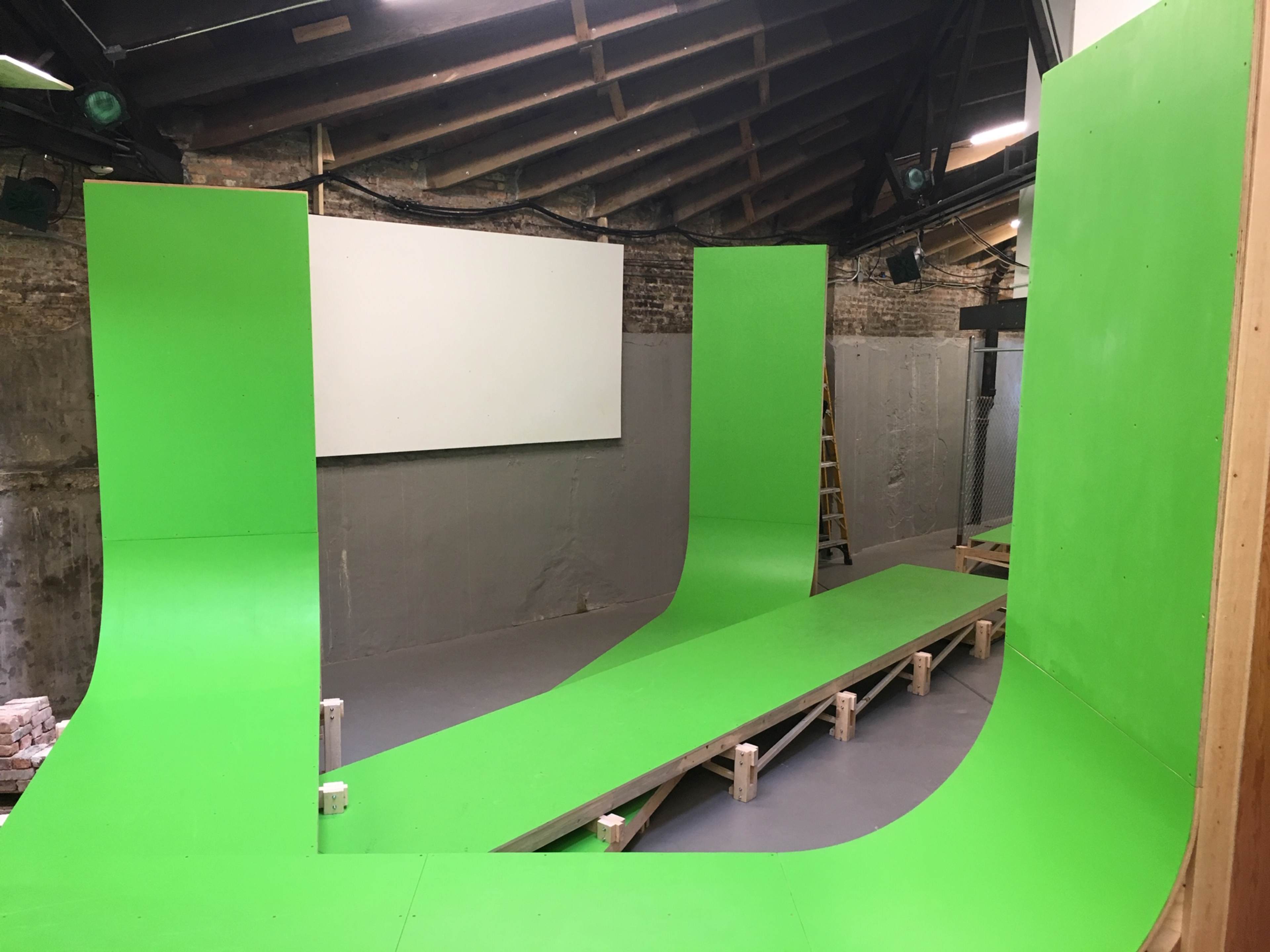 An indoor space with green ramps and a flat platform arranged in a curved layout against a backdrop of exposed brick walls and a blank screen.