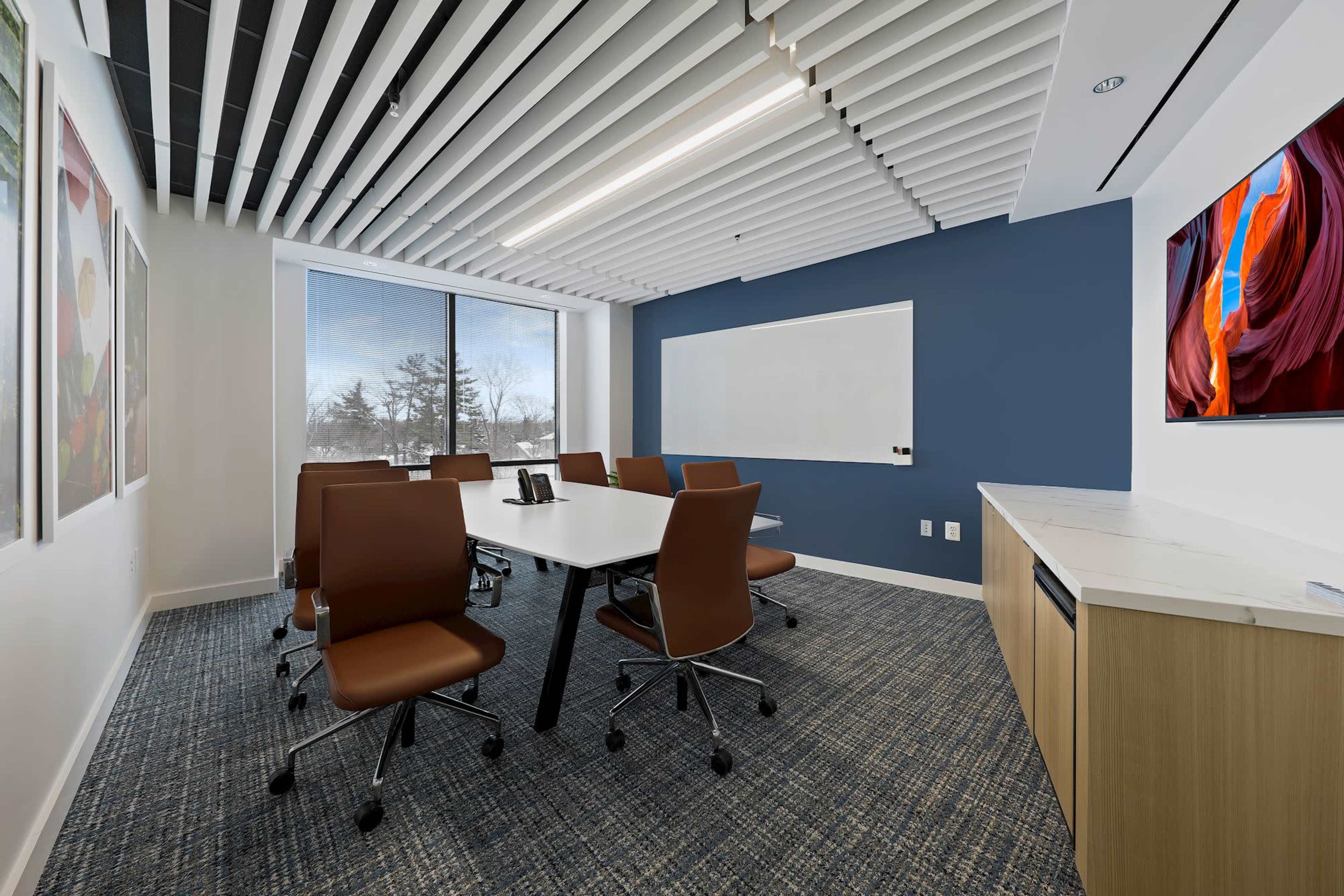 A modern conference room features a large table, ergonomic chairs, a whiteboard, and a wall-mounted TV, with large windows providing natural light.