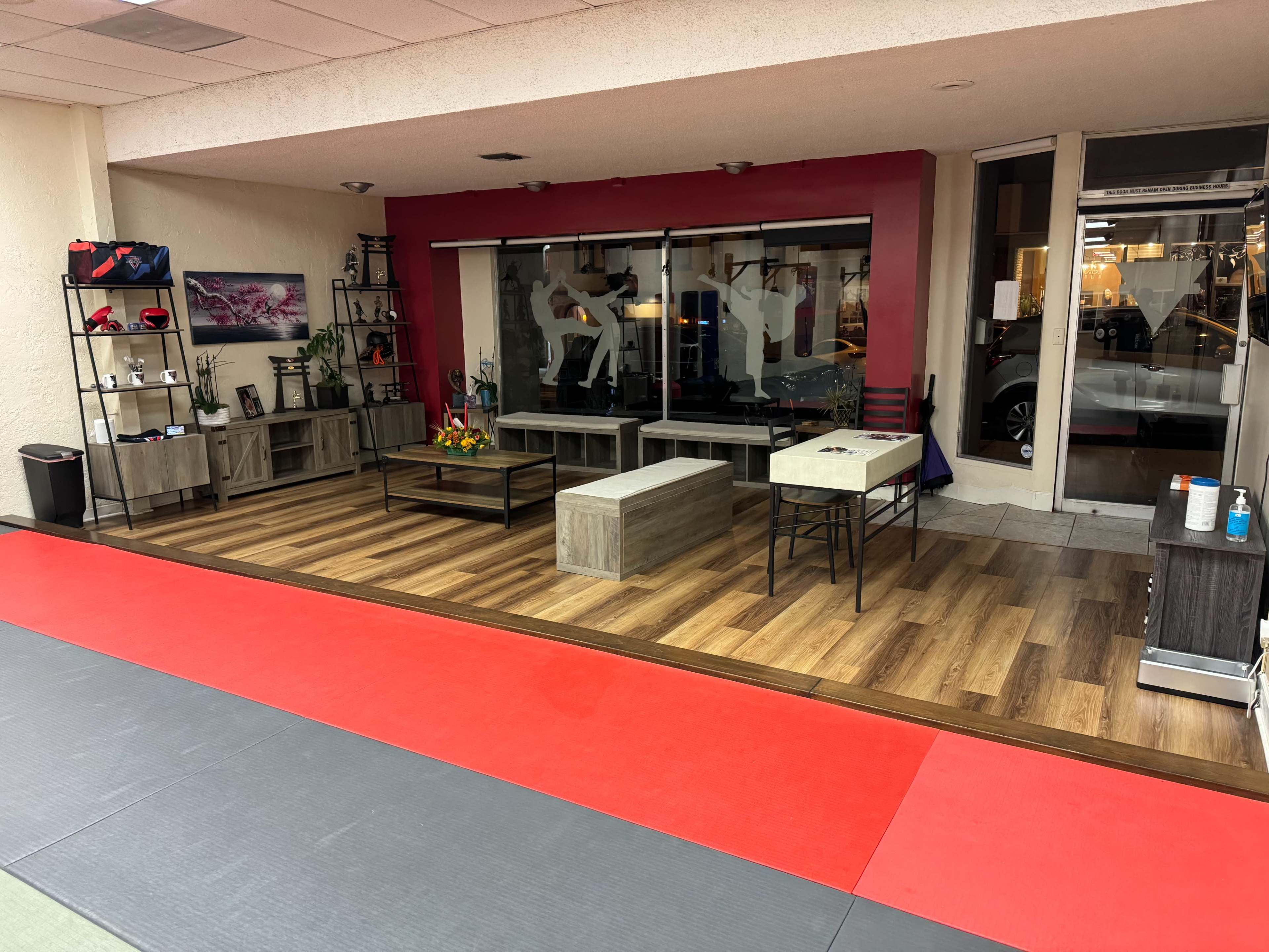 The interior of a martial arts studio features a red and wooden floor area with benches, decorative plants, and shelves displaying boxing equipment.