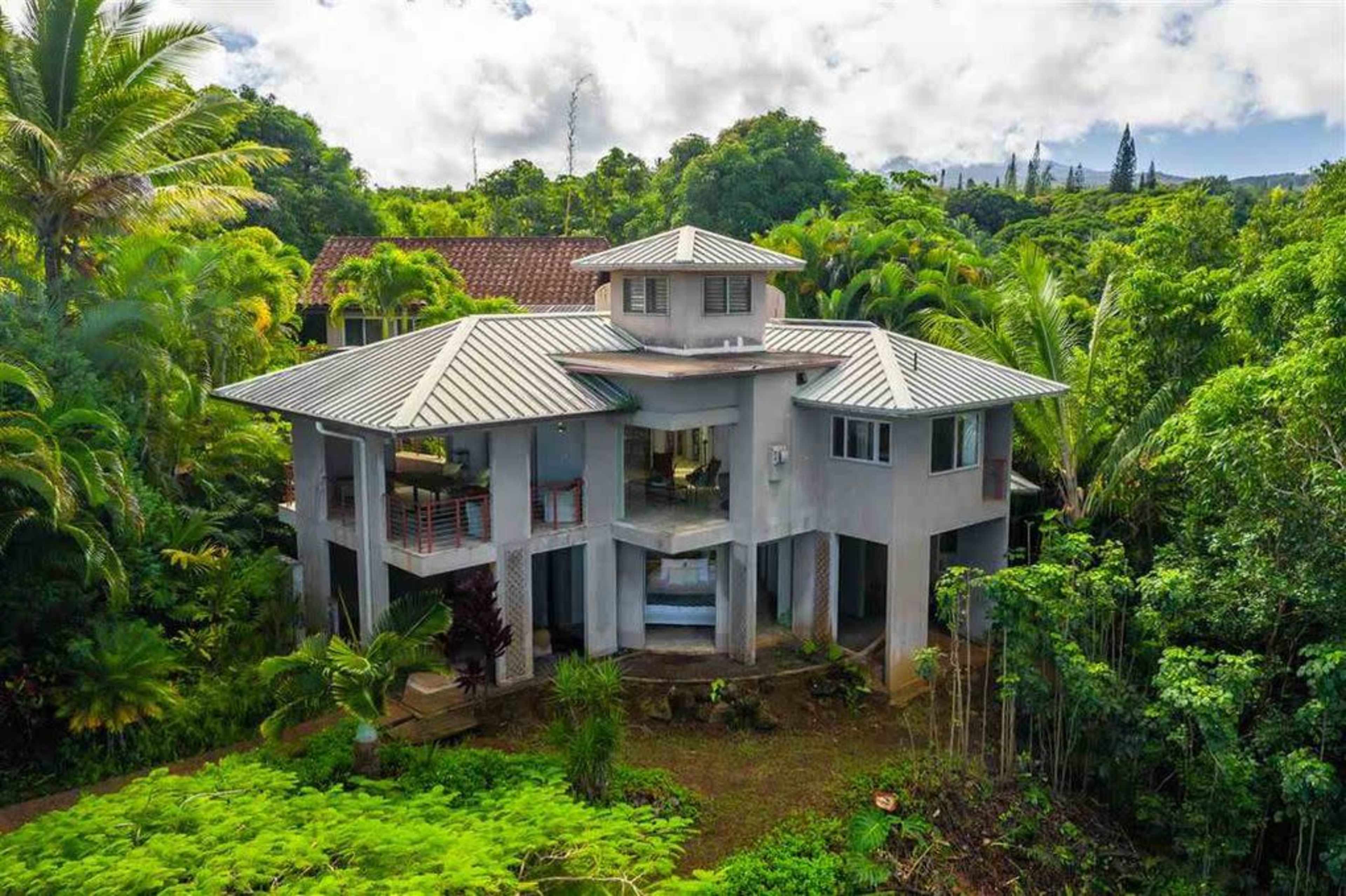 A modern, multi-level house surrounded by lush greenery and tall trees.