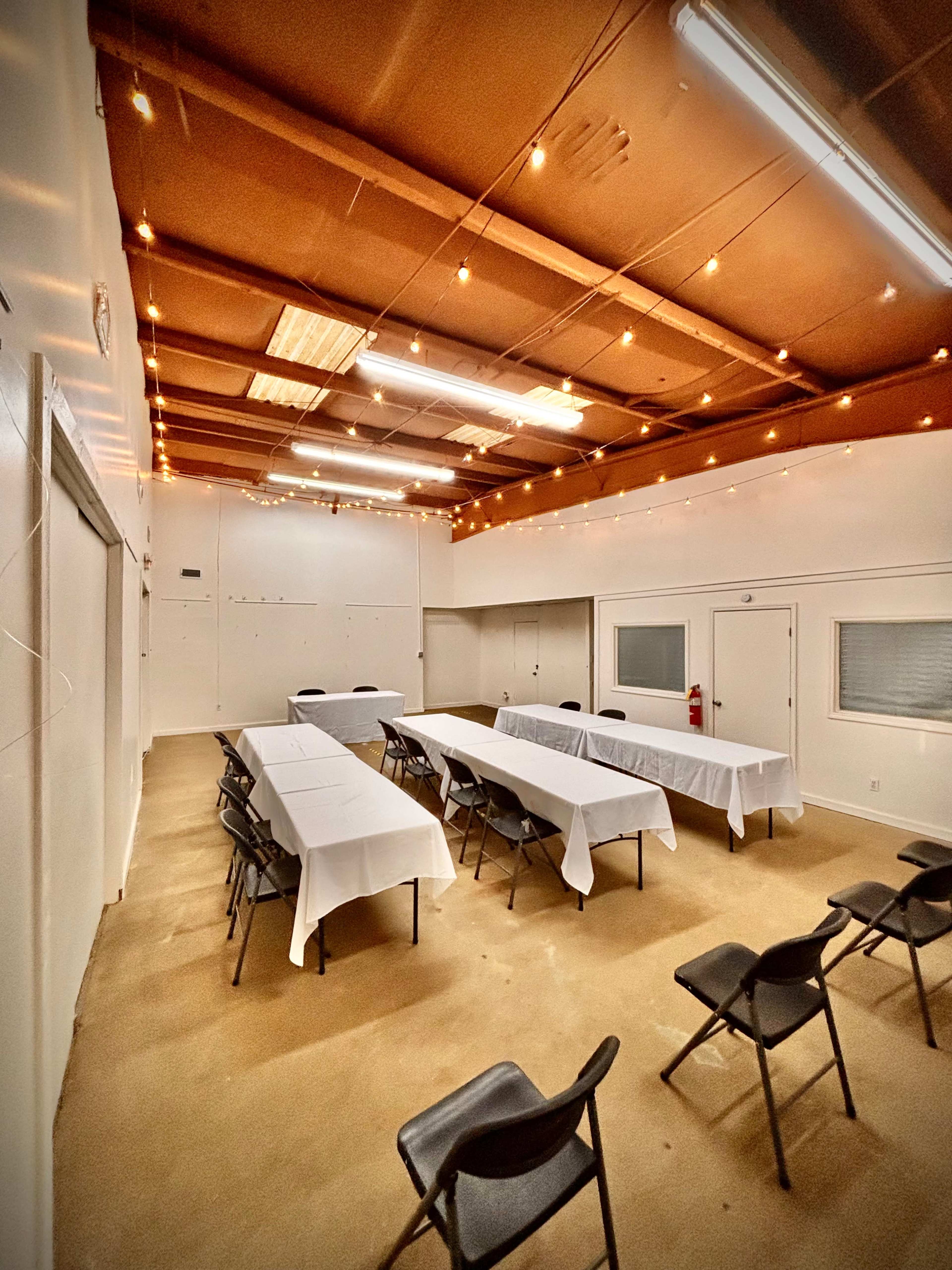 A spacious meeting room features several long tables covered with white tablecloths and black chairs arranged around them, illuminated by string lights on the ceiling.