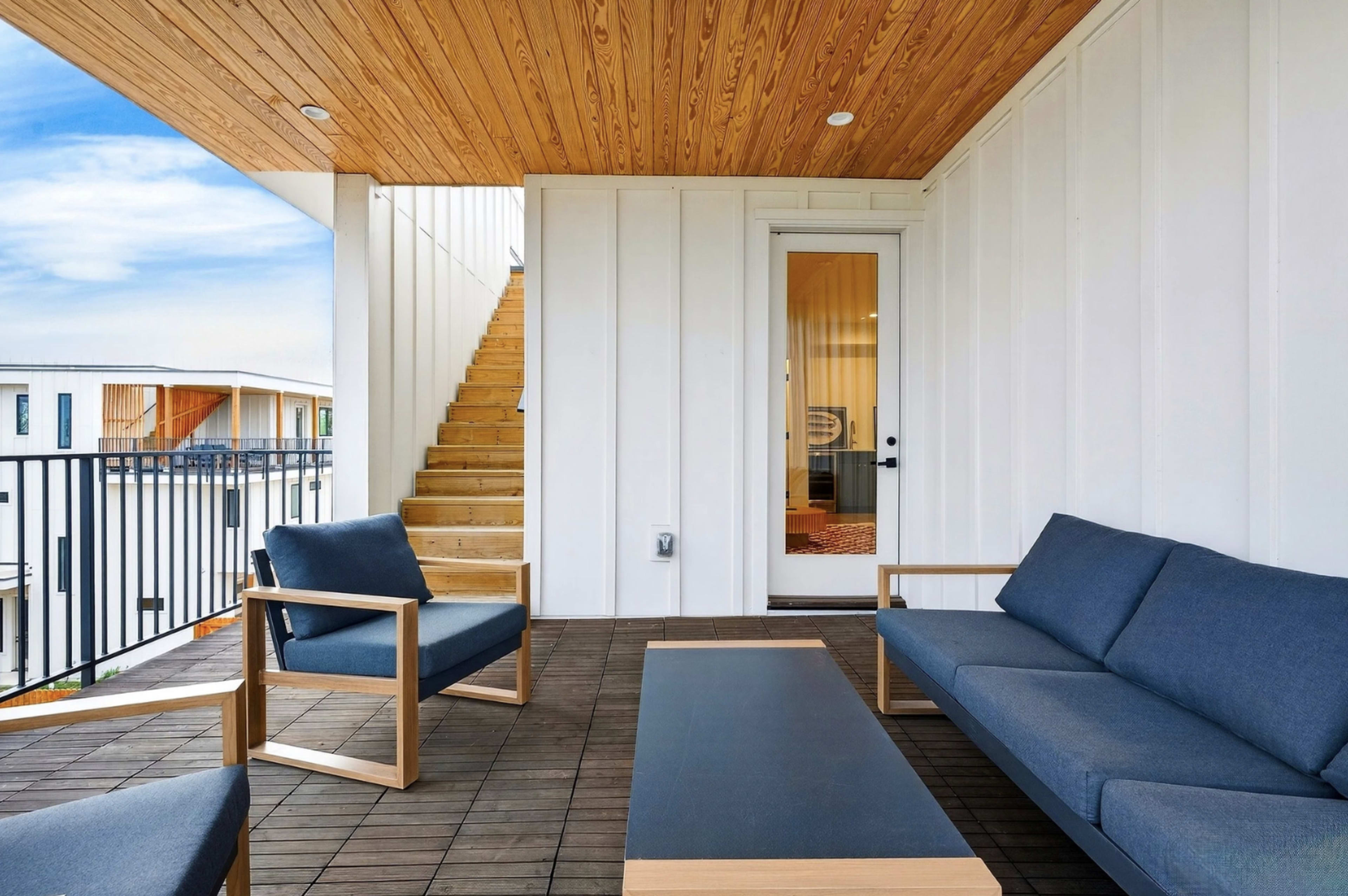 A modern balcony features wooden flooring, a minimalist seating area with blue cushions, and a staircase leading to an upper level.