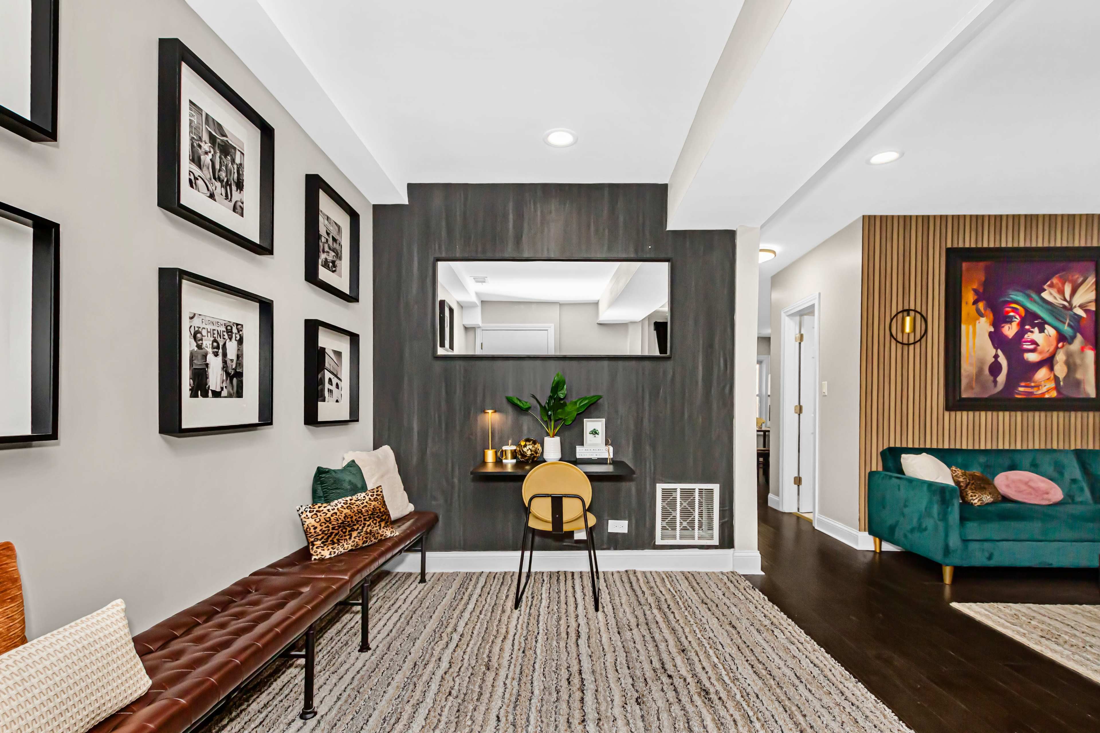 The scene features a living space with a bench, a small desk in front of a mirror, and framed photographs on the walls.