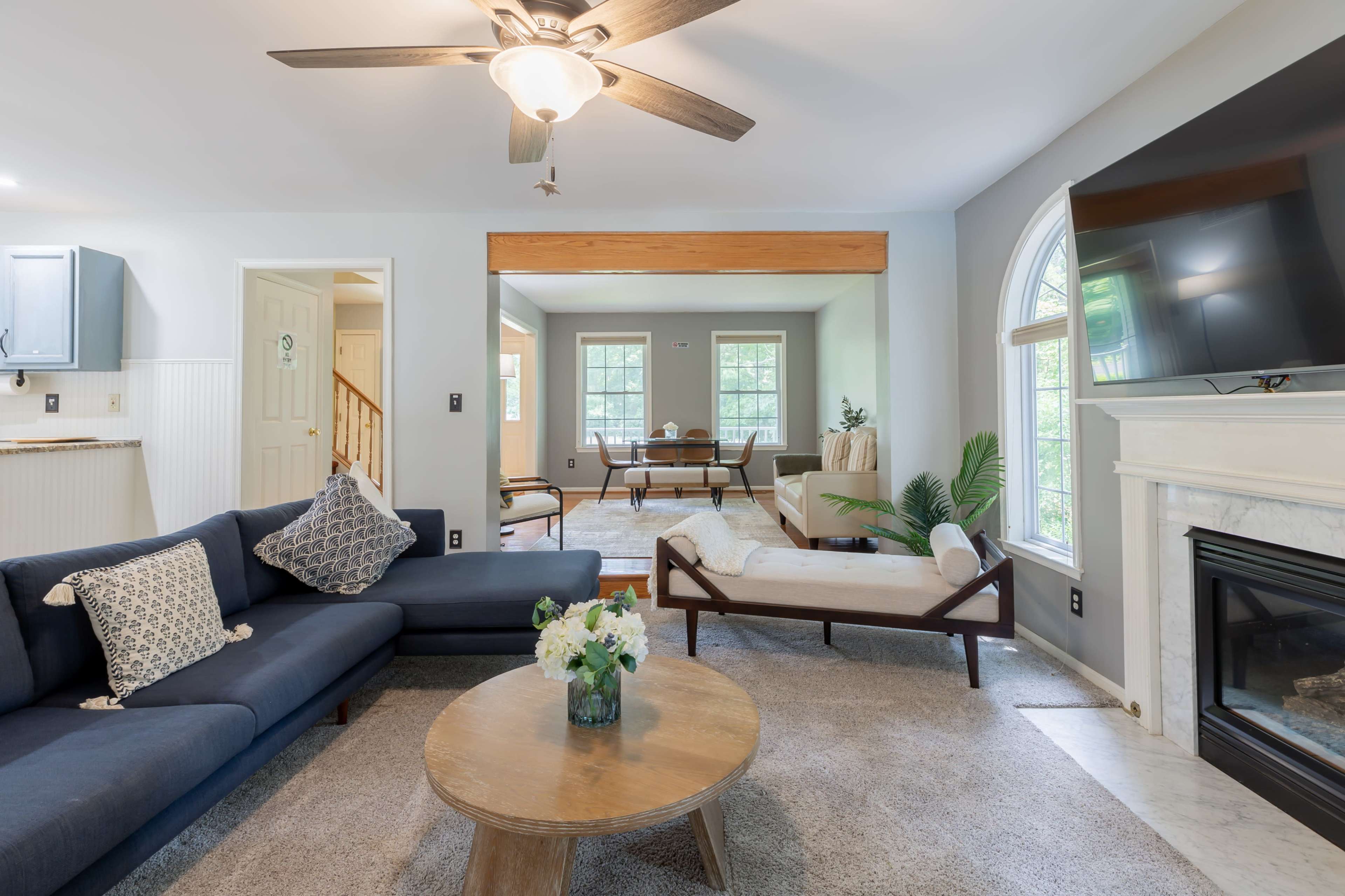A living room with a dark blue sectional sofa, a round coffee table, and large windows that provide natural light, leading to an adjacent dining area.