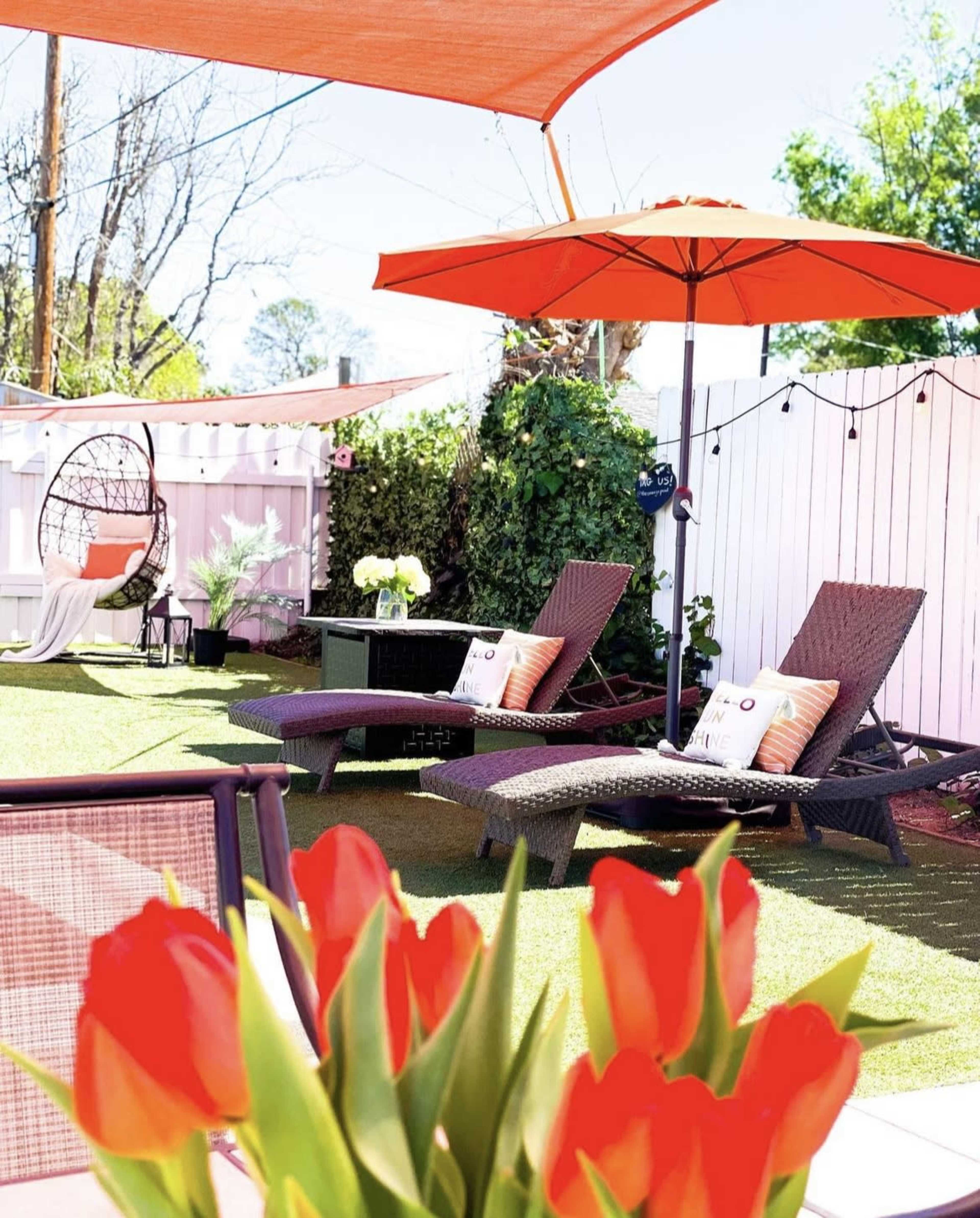 A patio area features two lounge chairs with pillows, an orange umbrella, a hanging chair, and vibrant red tulips in the foreground against a backdrop of greenery and a white fence.