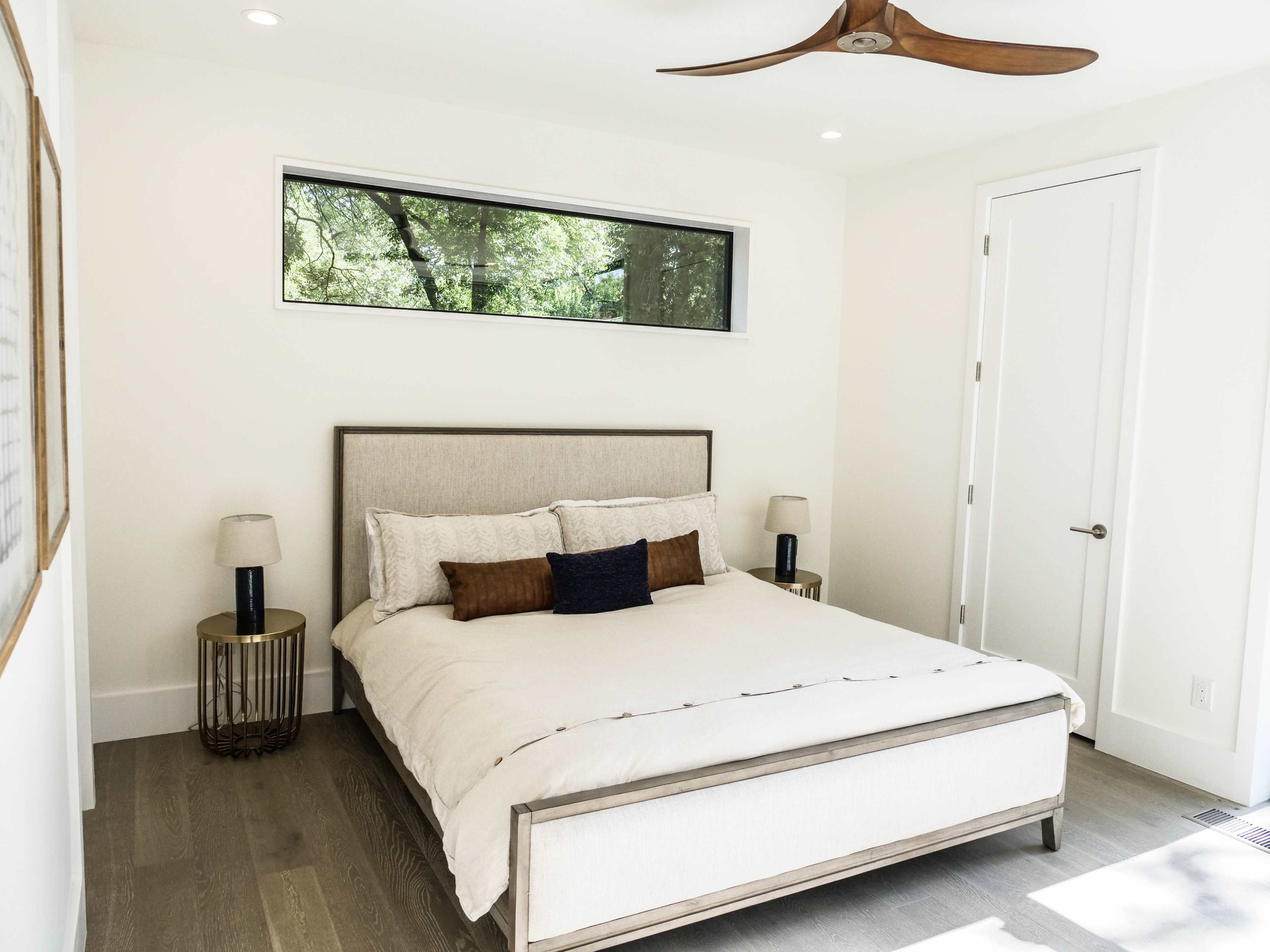A minimalist bedroom features a queen-sized bed with light bedding, two side tables, and a large window showcasing green trees outside.