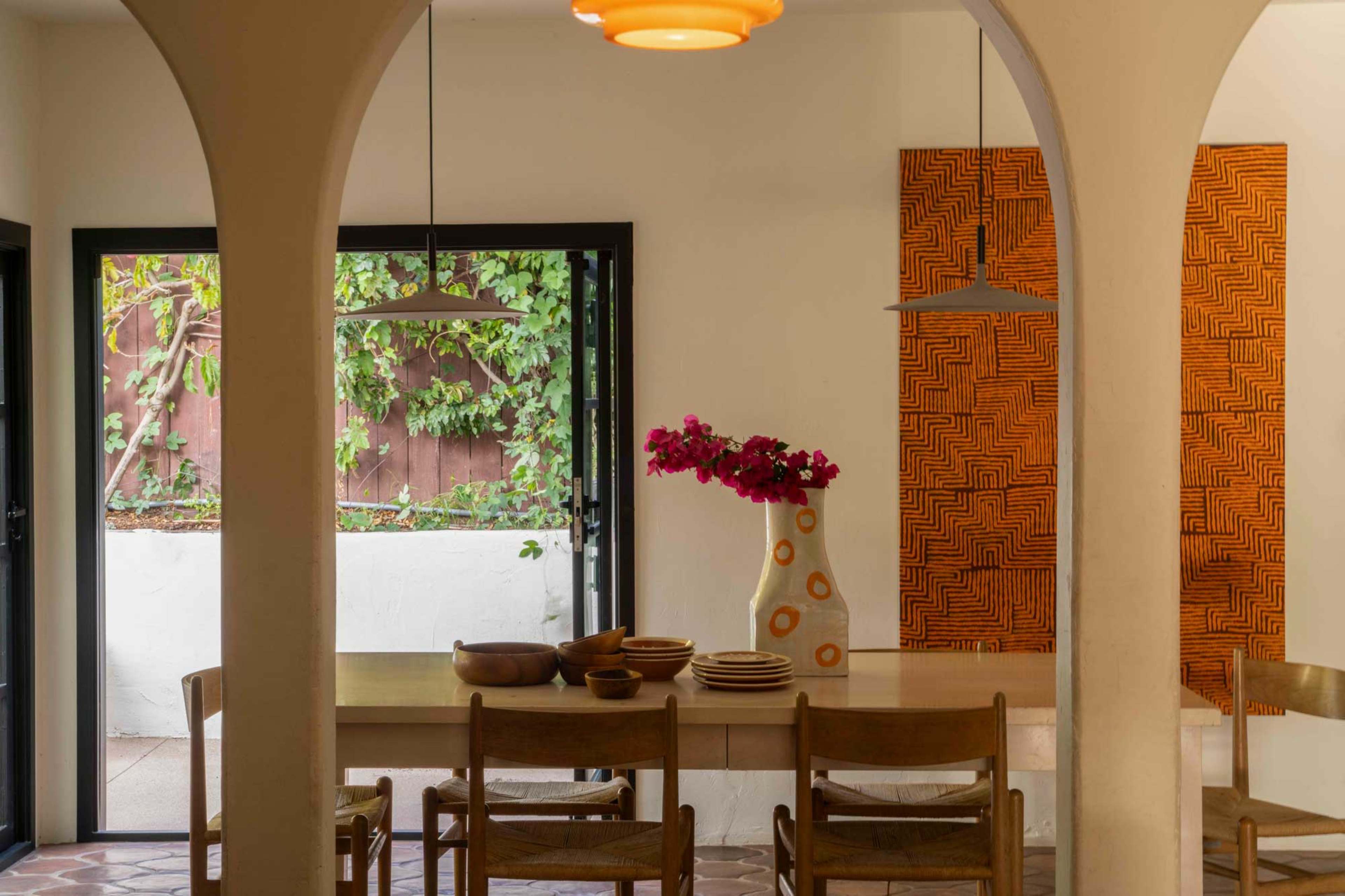 A dining area features a long table with bowls and plates, surrounded by wooden chairs, with a view of a garden visible through an arched doorway.