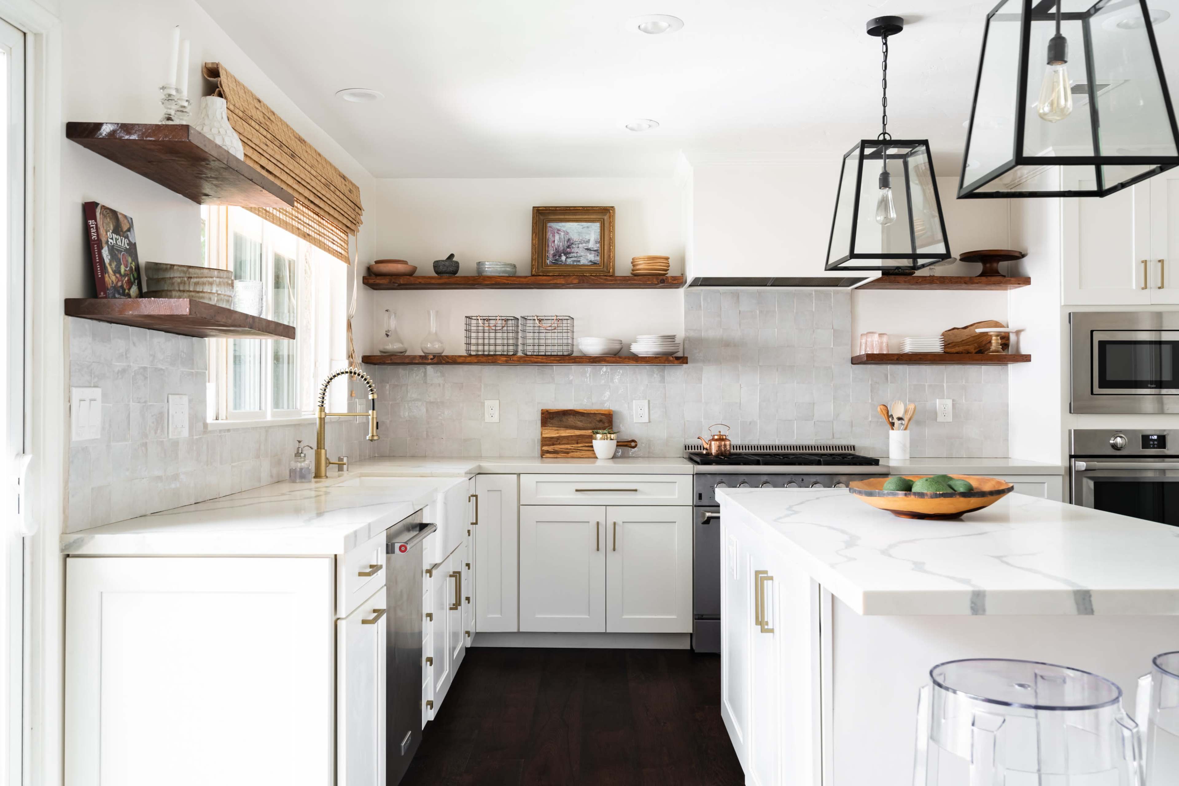 A modern kitchen features white cabinetry, marble countertops, open shelving with decorative items, and stainless steel appliances.