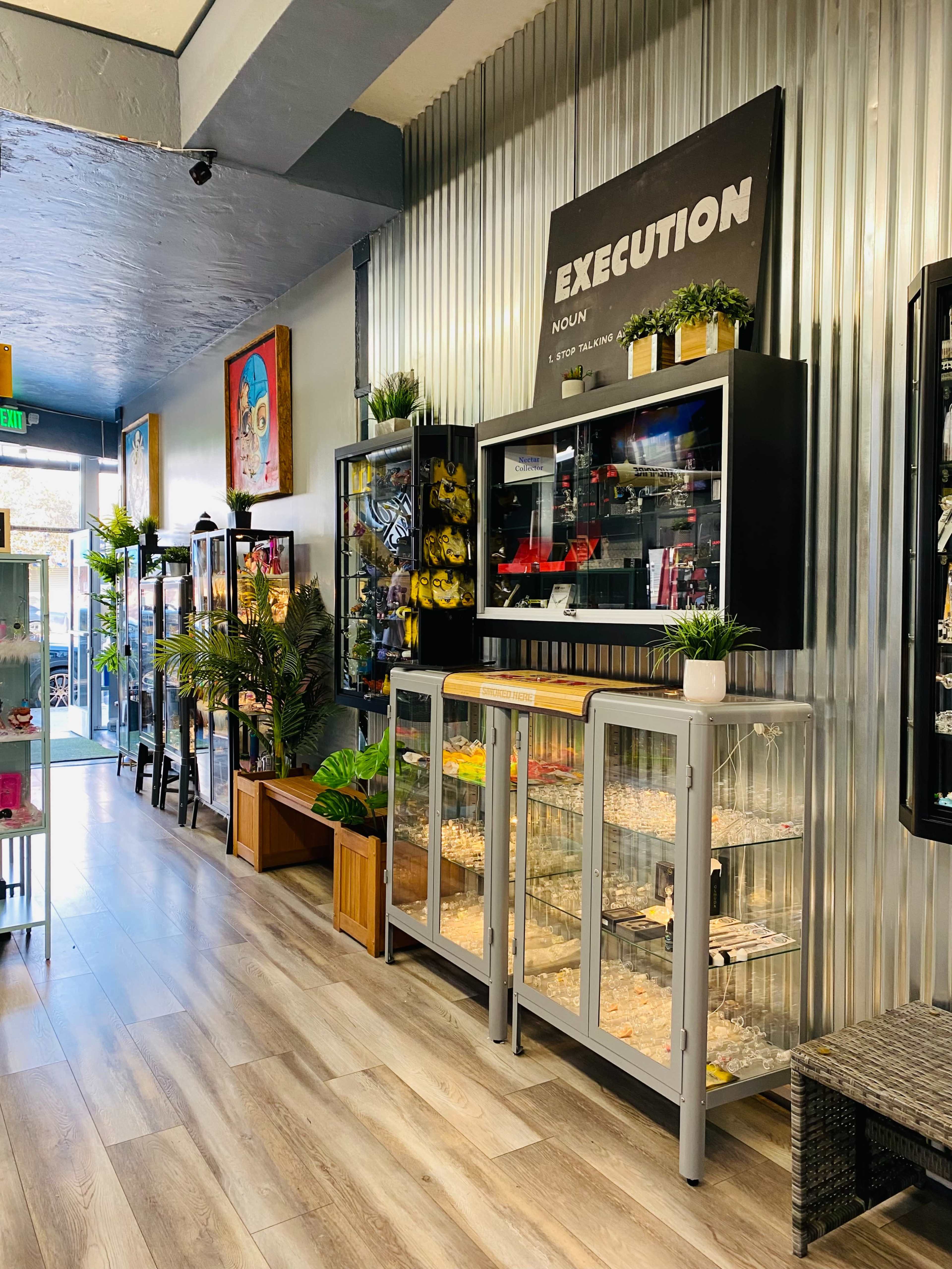 A narrow retail space with glass display cases containing various items, framed artwork, and potted plants along a wall that features a sign labeled "EXECUTION."