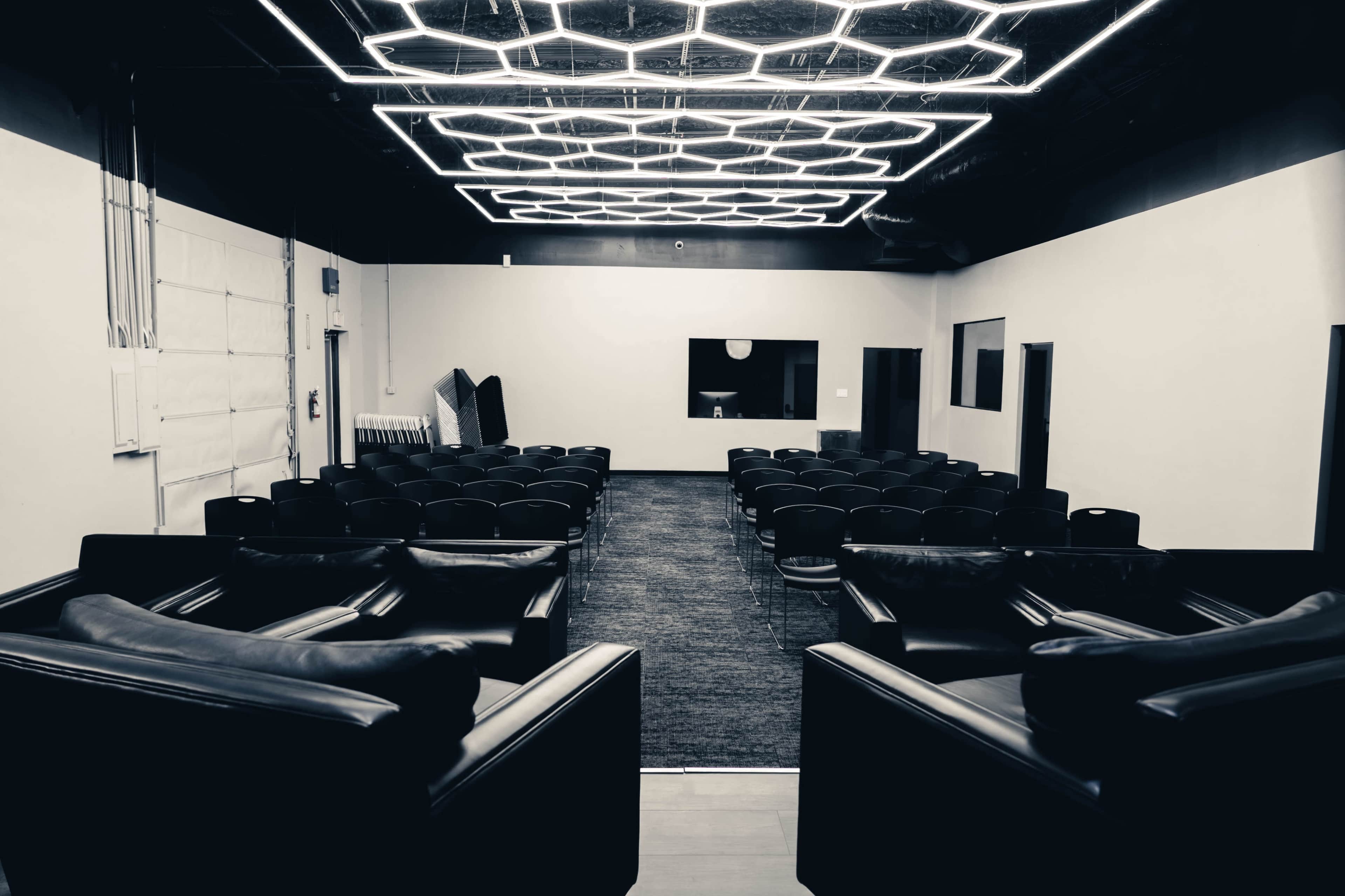 The image shows a modern, sparsely furnished indoor space designed for presentations, featuring rows of chairs facing a blank wall with a screen, in front of a large, illuminated ceiling fixture.