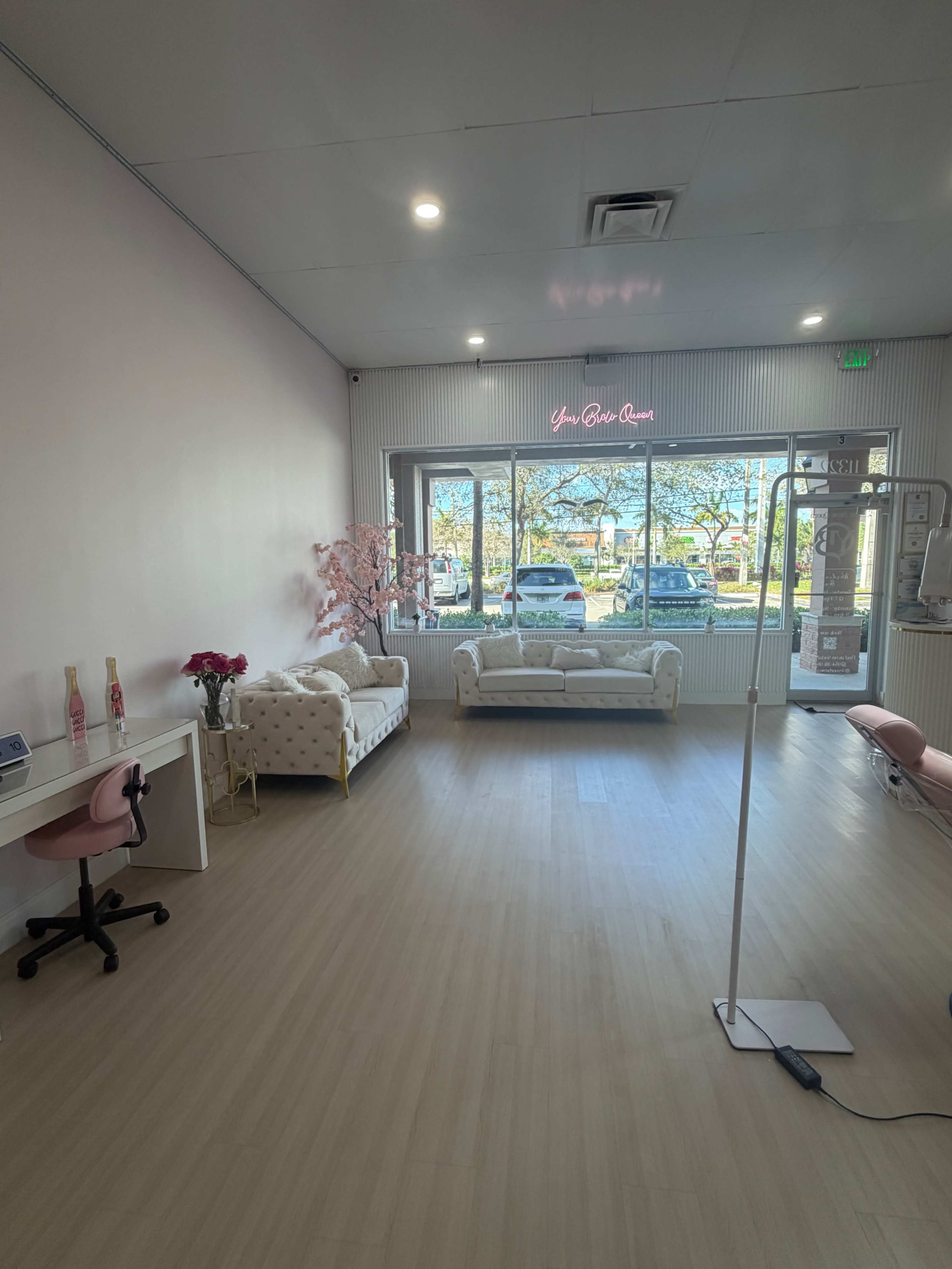 The image shows a bright, modern salon interior with two white sofas, a small table with flowers, and large windows letting in natural light.